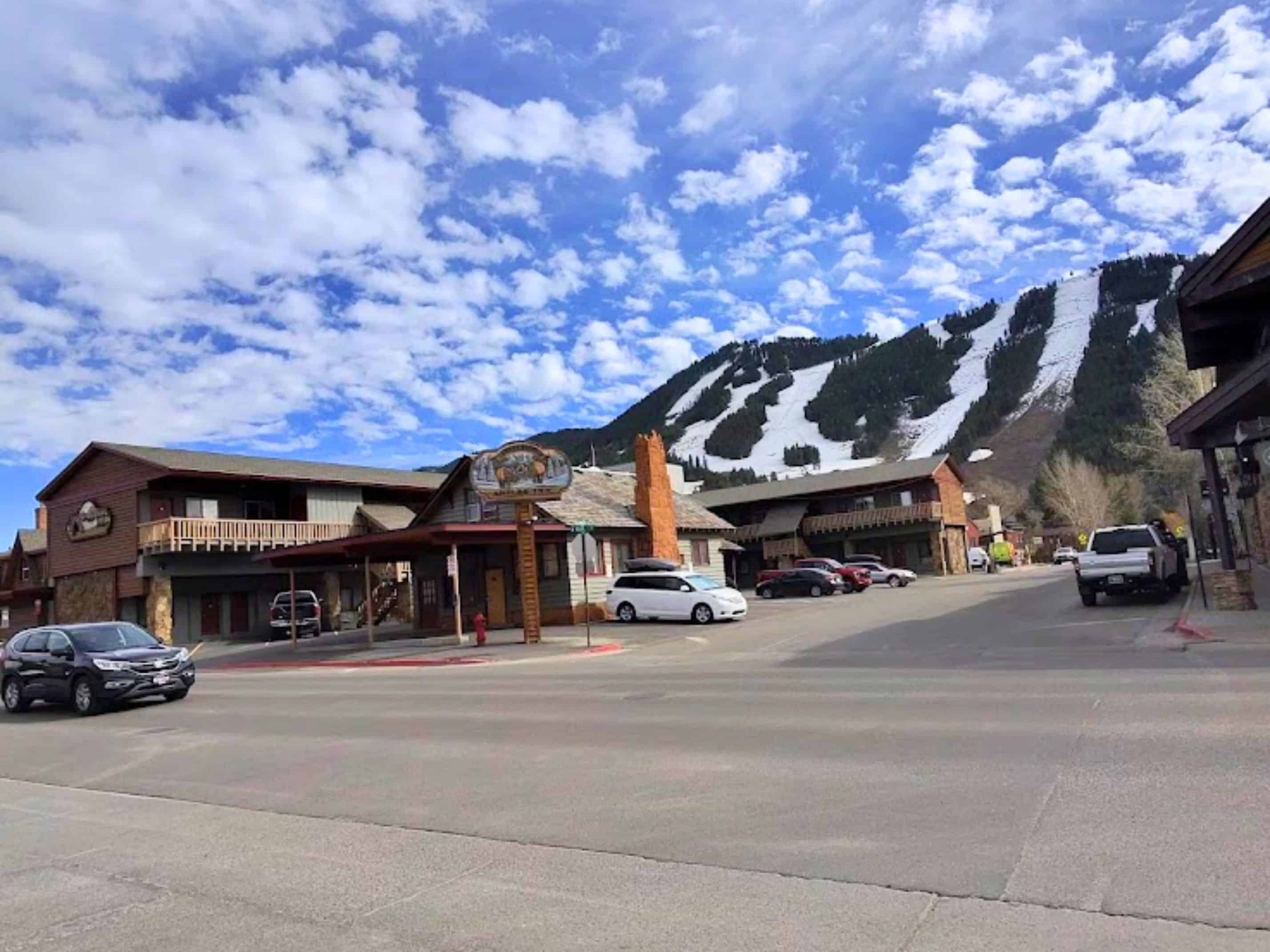 Cars parked near rustic wooden buildings with a mountain and ski slopes in the background under a cloudy sky&mdash;this scene perfectly captures the charm highlighted in any Jackson Hole travel guide.
