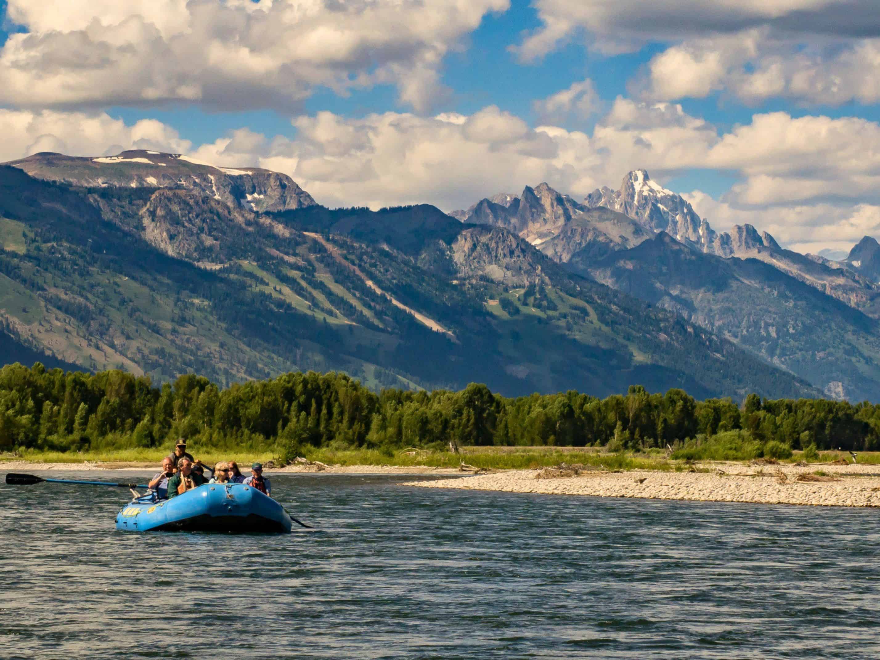 People rafting on a river with forested banks and tall, rugged mountains under a partly cloudy sky&mdash;a scene straight out of a Jackson Hole travel guide.