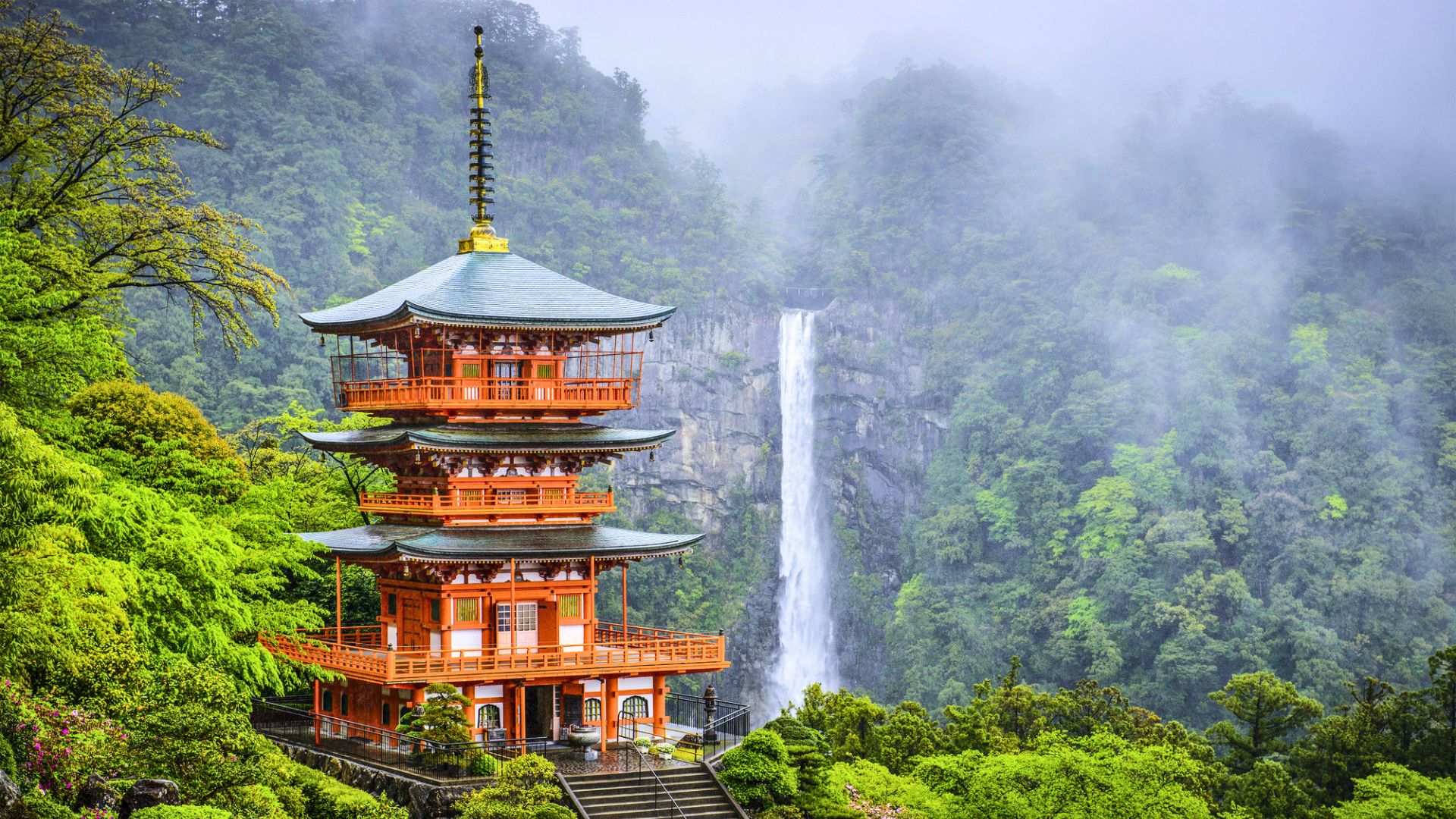 A red pagoda stands amid lush greenery with a tall waterfall cascading down in the misty background, capturing the serene beauty often discovered while traveling in Japan.