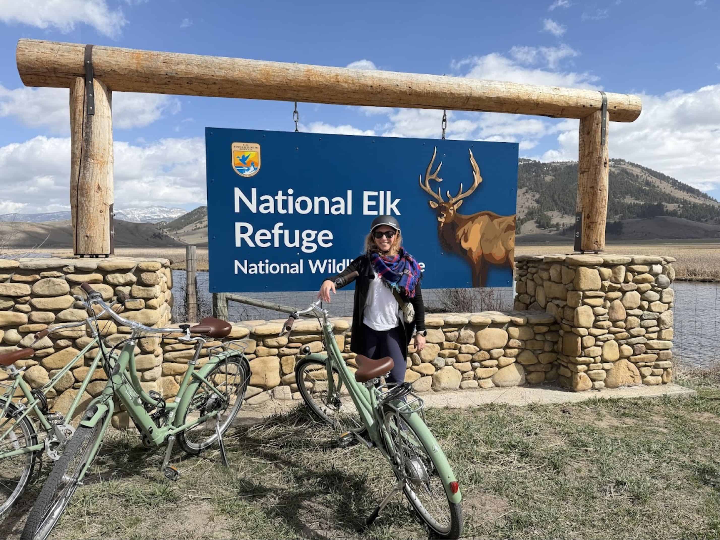 Person with a bike stands by a "National Elk Refuge" sign; mountains and blue sky in the background, capturing a classic scene from any Jackson Hole travel guide.