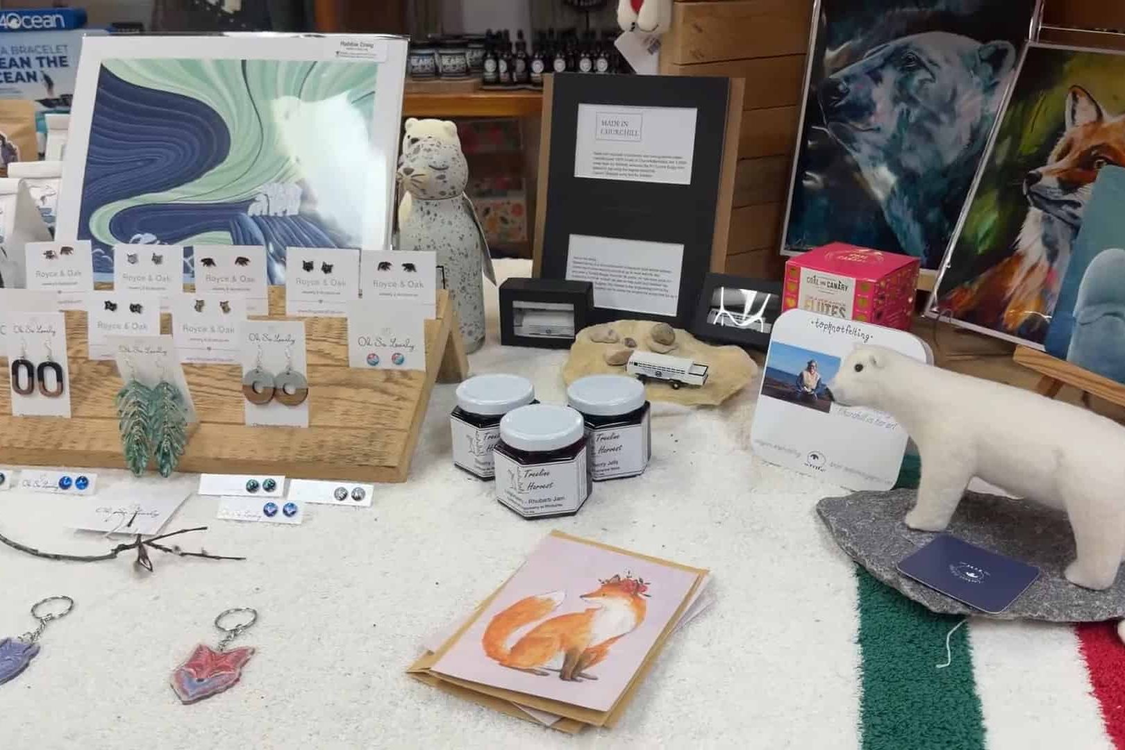 Gift shop display with animal art, jewelry, cards, keychains, candles, and a plush polar bear on a table&mdash;perfect souvenirs for your Churchill Northern Lights tour.