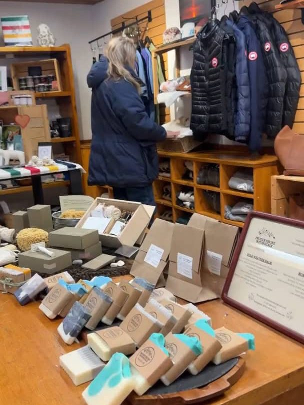 A woman browses shelves in a shop selling soaps, clothes, and various goods displayed on wooden tables, reminiscent of the unique treasures one might discover after a day on a Churchill Northern Lights tour.