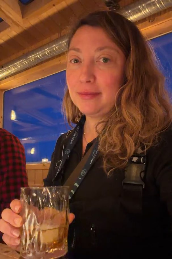 Woman with wavy hair holding a drink, sitting at a wooden table inside a cozy, warmly lit room, reminiscing about her unforgettable Churchill Northern Lights tour.
