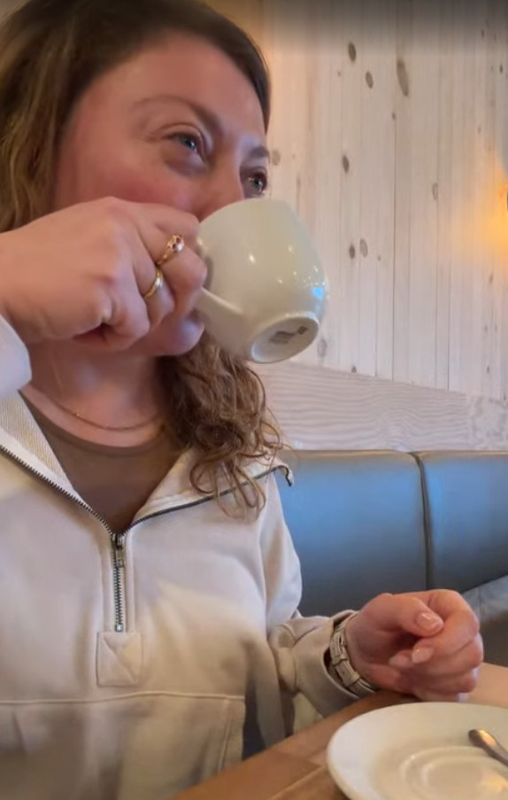 Woman in a cream jacket drinks from a white cup in a cozy, wood-paneled caf&eacute;, perhaps sharing stories from her recent Churchill Northern Lights tour.