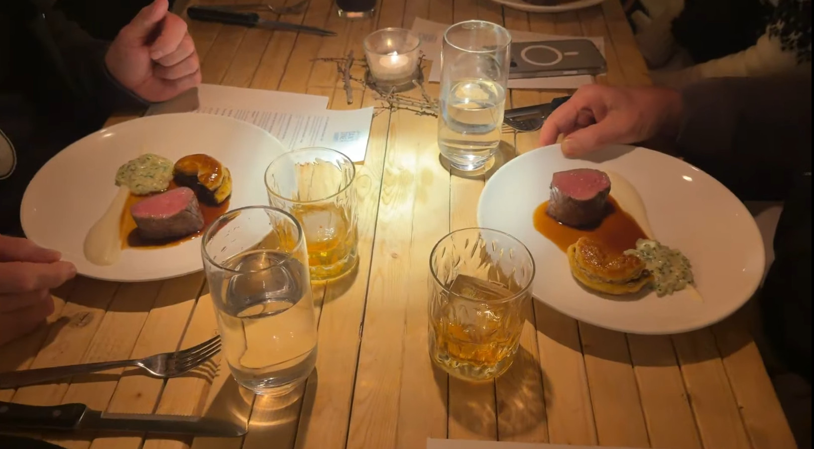Two plates with steak, sauce, and garnish on a wooden table with drinks, water glasses, and candles&mdash;an inviting scene reminiscent of a memorable evening after a Churchill Northern Lights tour.