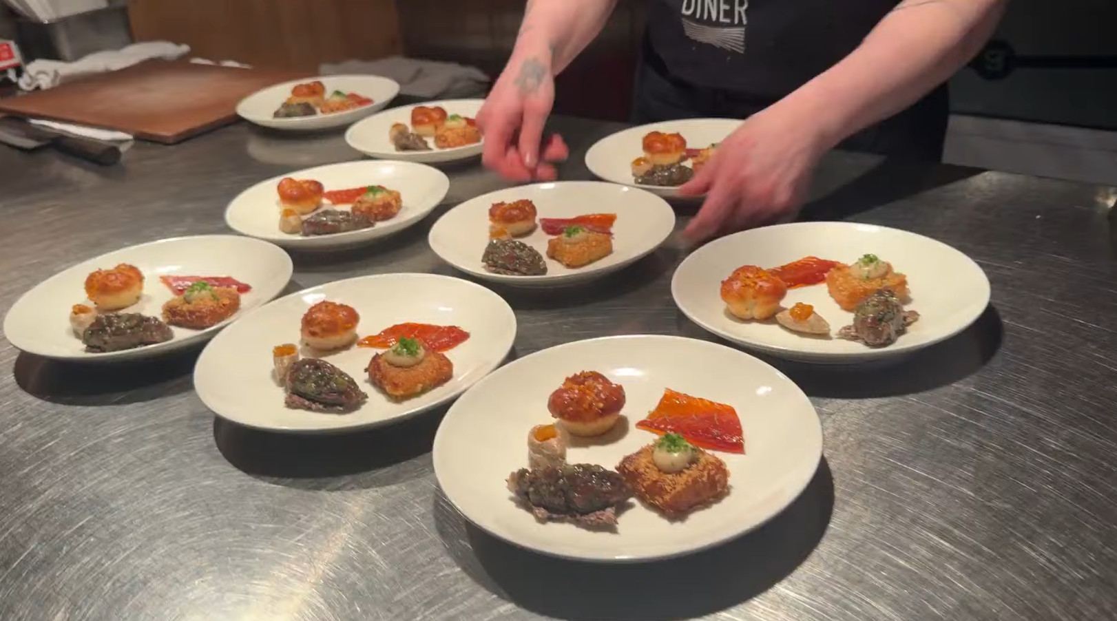 A person arranges plates of gourmet appetizers on a metal counter in a restaurant kitchen, reminiscent of the meticulous care found after a long day spent on a Churchill Northern Lights tour.