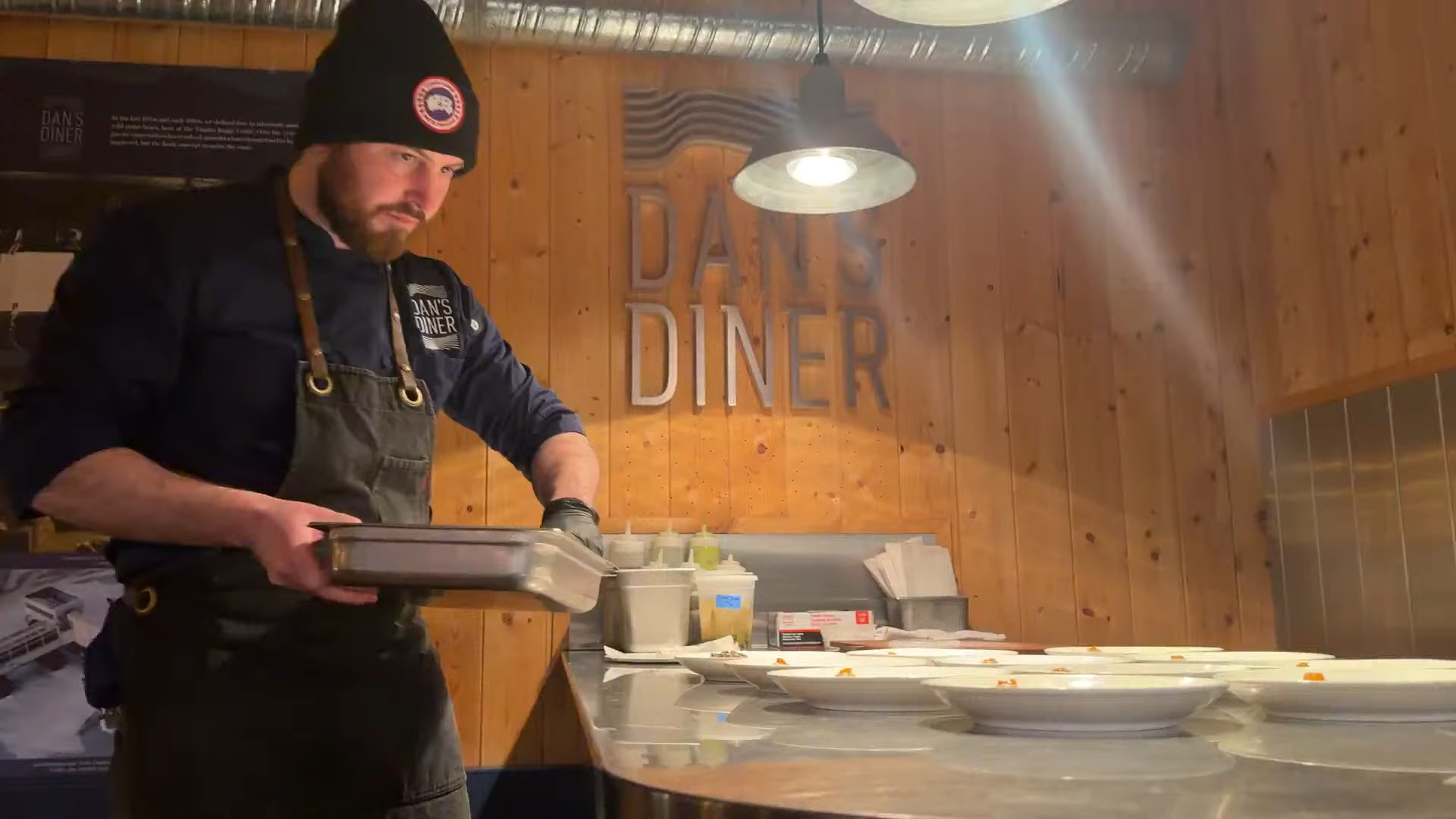 Chef plating food in a wooden diner kitchen, wearing a beanie and apron, under hanging lights&mdash;capturing the cozy spirit of a Churchill Northern Lights tour.