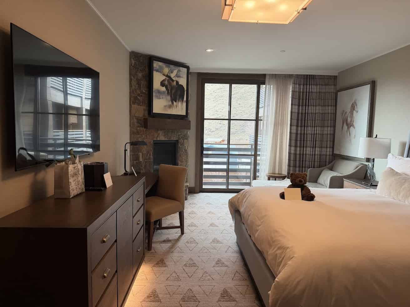 Modern hotel room with a bed, TV, desk, chair, and large window with view; teddy bear on bed&mdash;an inviting retreat for any Jackson Hole travel guide adventure.