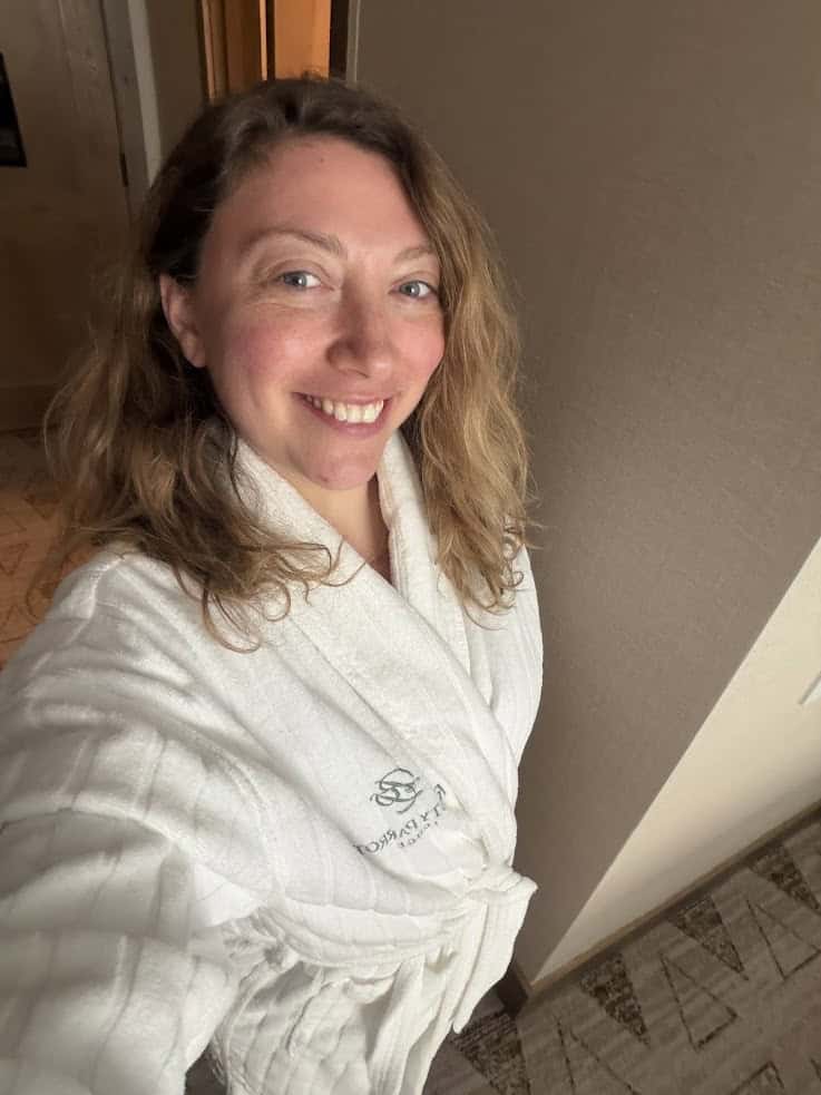 Woman with wavy hair smiles, wearing a white robe in a warmly lit indoor setting&mdash;her relaxed demeanor reminiscent of the serene moments found in any great Jackson Hole travel guide.