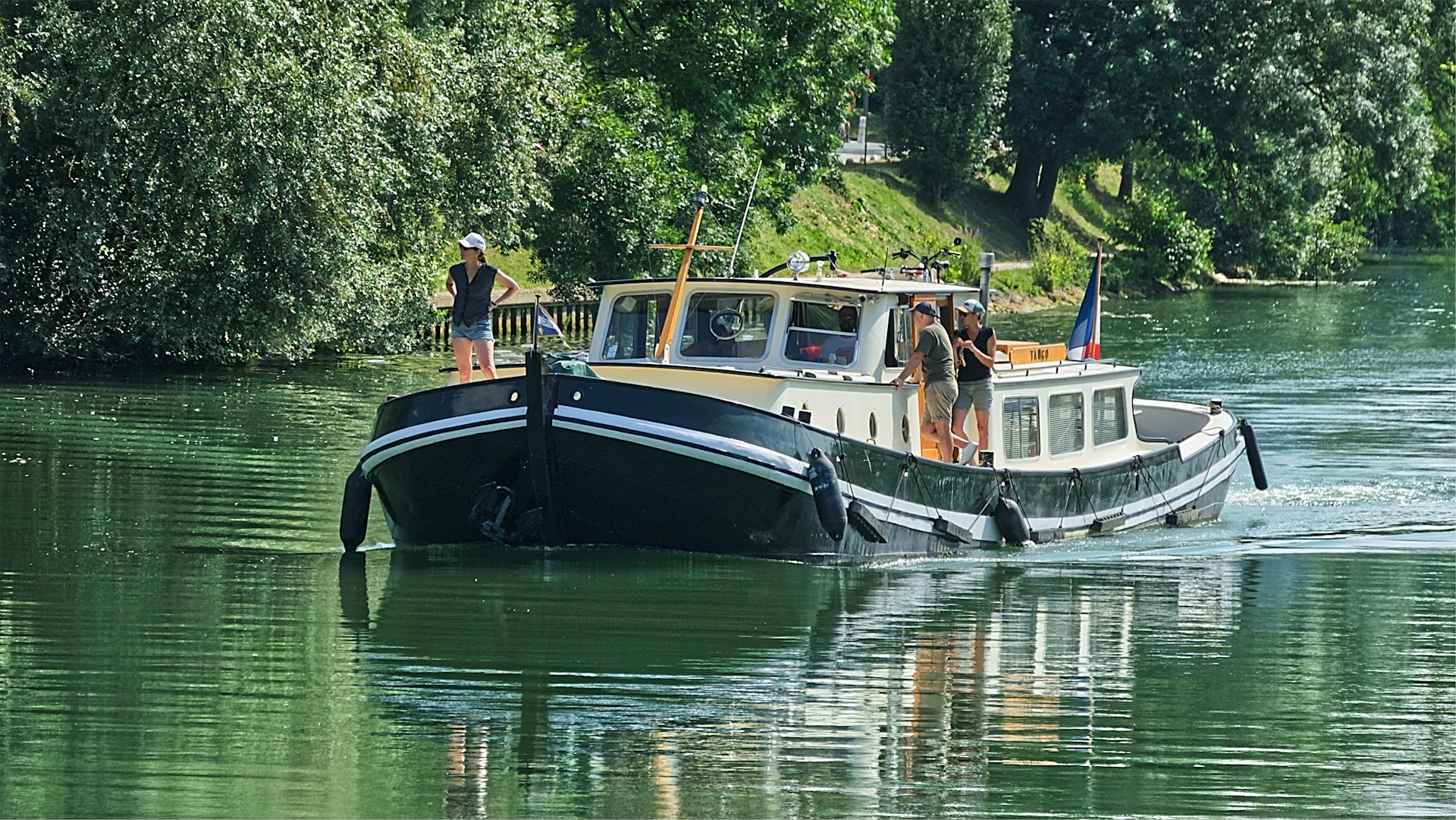 A black and white boat, reminiscent of a luxury barge cruise, with three people aboard travels on a calm, green river surrounded by trees.
