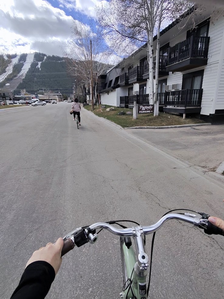 View from a bike following another cyclist on a street beside a white building, with mountains in the distance&mdash;a scene right out of a Jackson Hole travel guide.