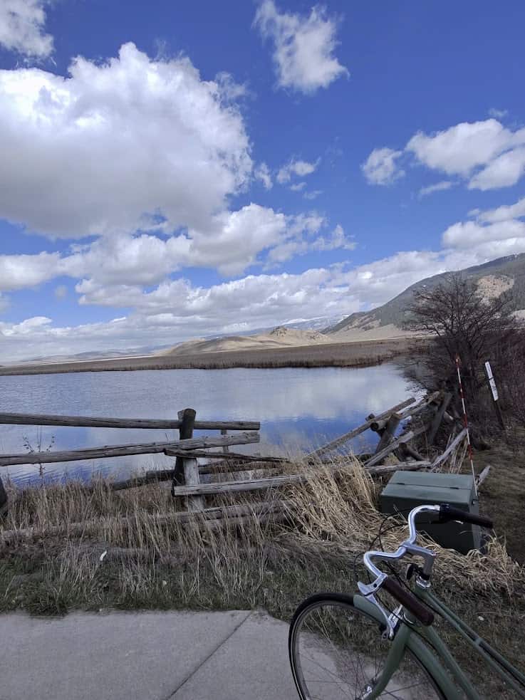 A lake with mountains and clouds, grassy shore, wooden fence, and part of a bicycle in the foreground&mdash;this idyllic scene could be straight out of a Jackson Hole travel guide.