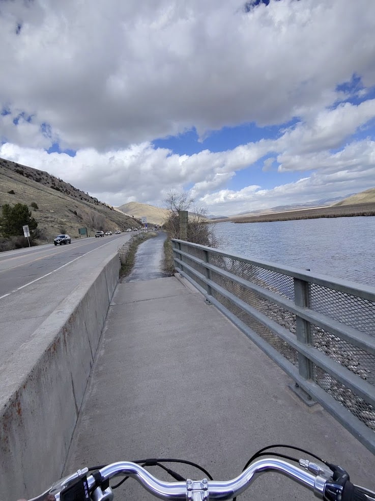 View from a bike on a riverside path, with a road and hills on the left under a cloudy sky&mdash;a scene reminiscent of journeys featured in a Jackson Hole travel guide.
