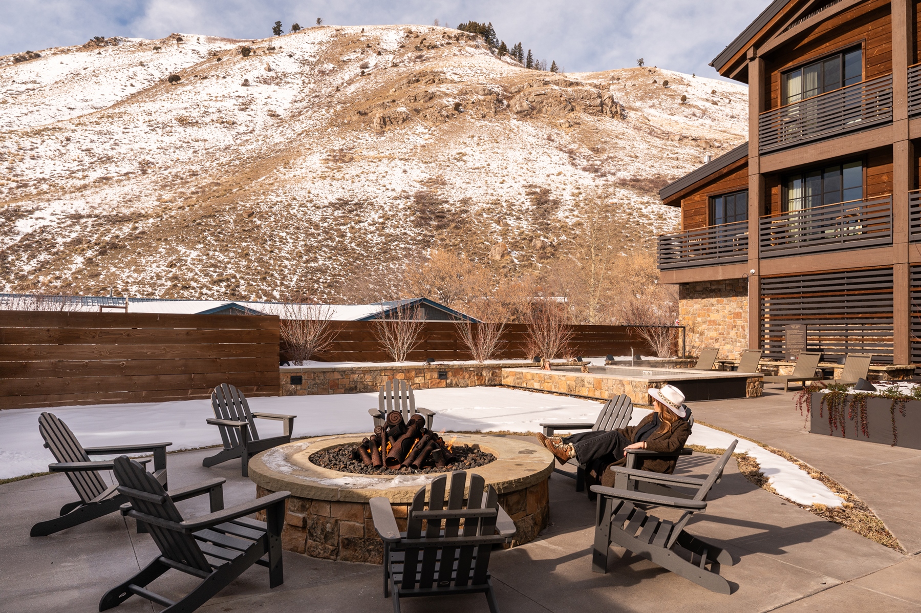 A person sits by a fire pit in a patio area at the Rusty Parrot Lodge and Spa, with snowy mountains and the inviting lodge in the background.