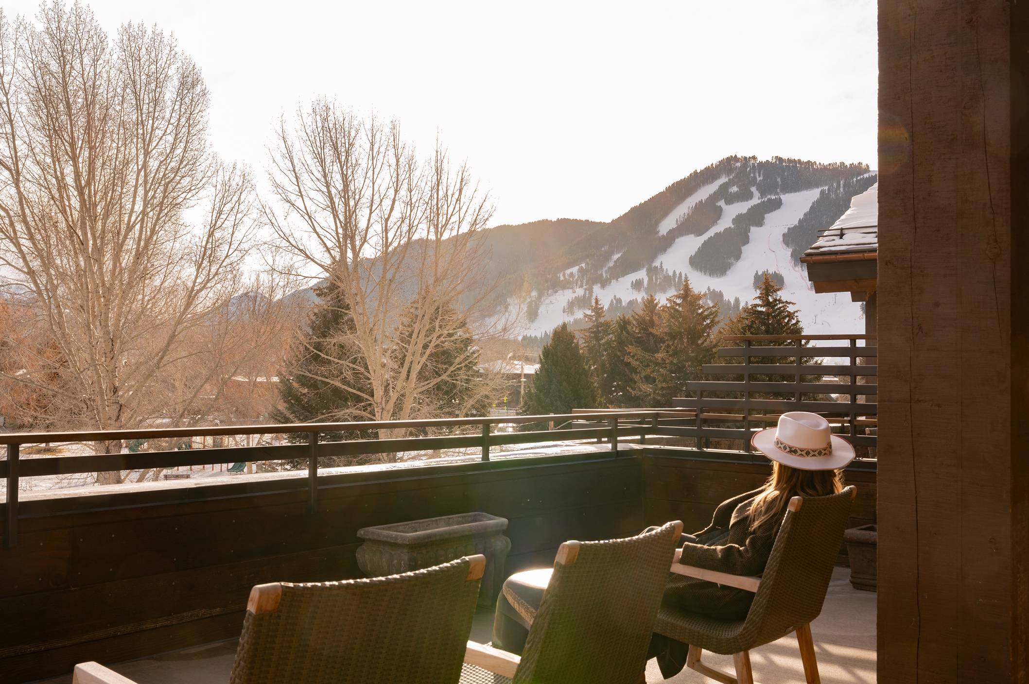Woman in a white hat sits on a balcony at Rusty Parrot Lodge and Spa, overlooking snowy mountains and bare trees in sunlight.