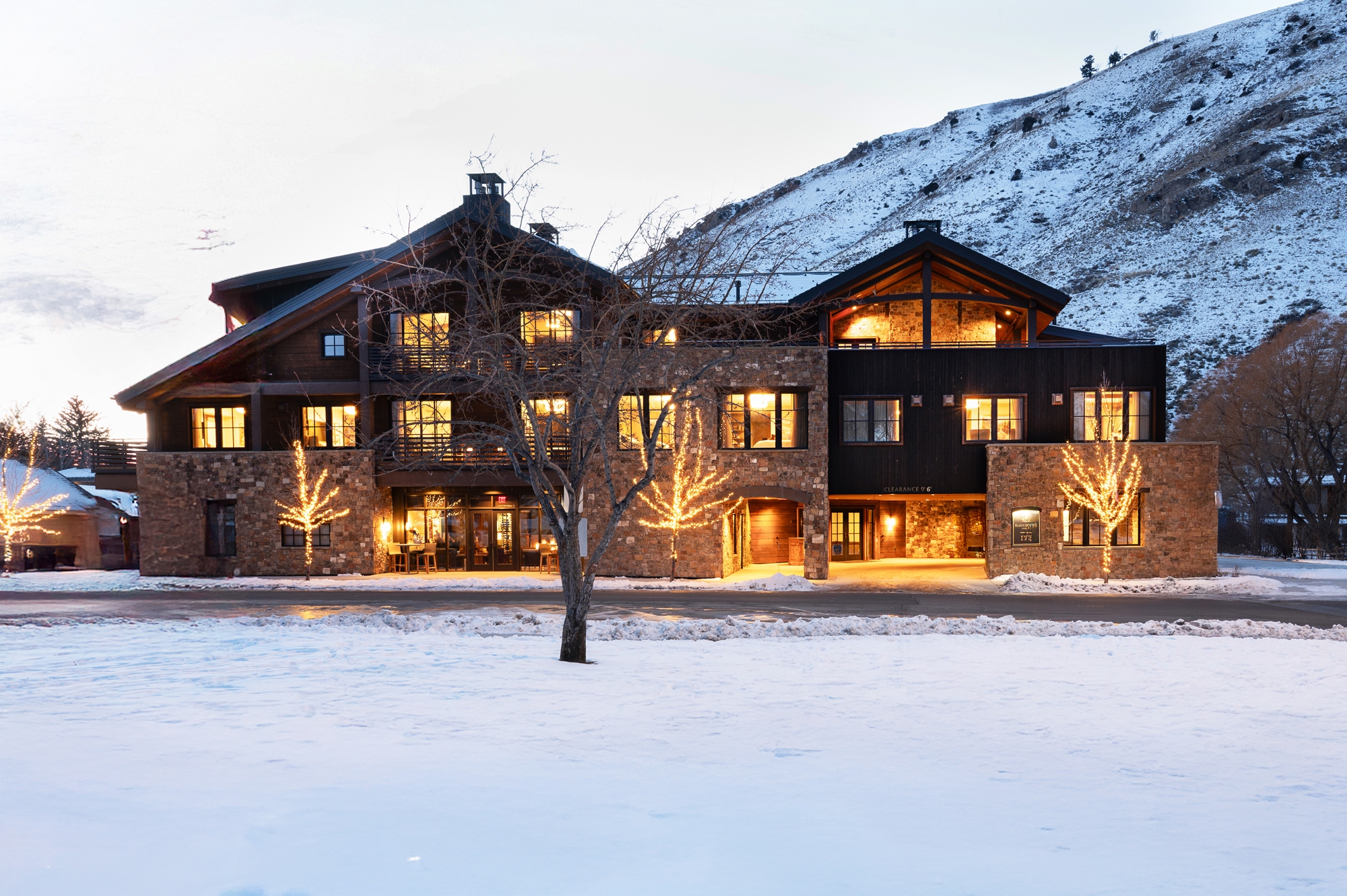A large lodge with glowing lights, set against a snowy landscape and a hill at dusk, reminiscent of the cozy elegance found at the Rusty Parrot Lodge and Spa.