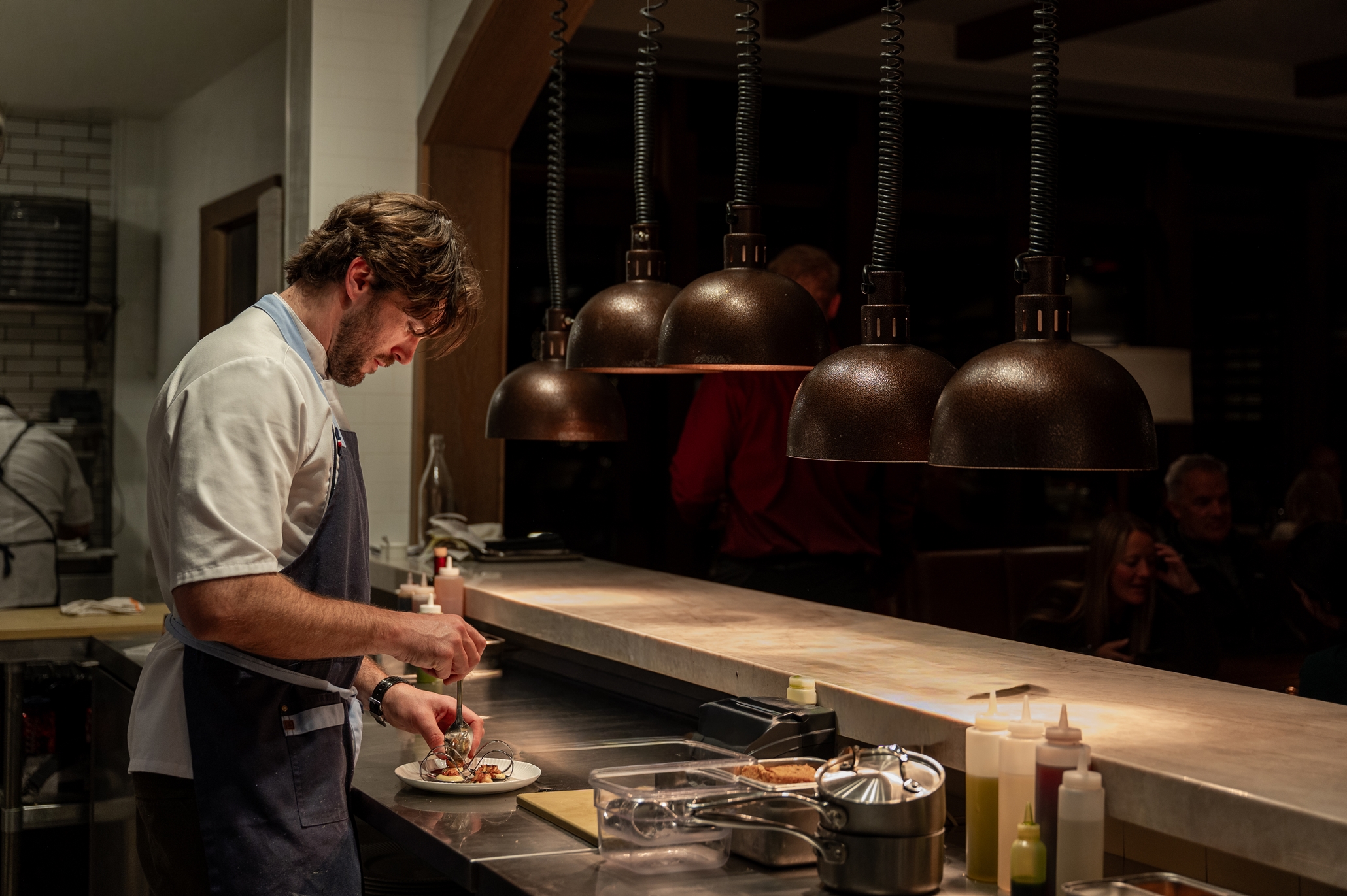 Chef plating food in a dimly lit kitchen at the Rusty Parrot Lodge and Spa, with bottles and utensils on the counter under hanging lamps.