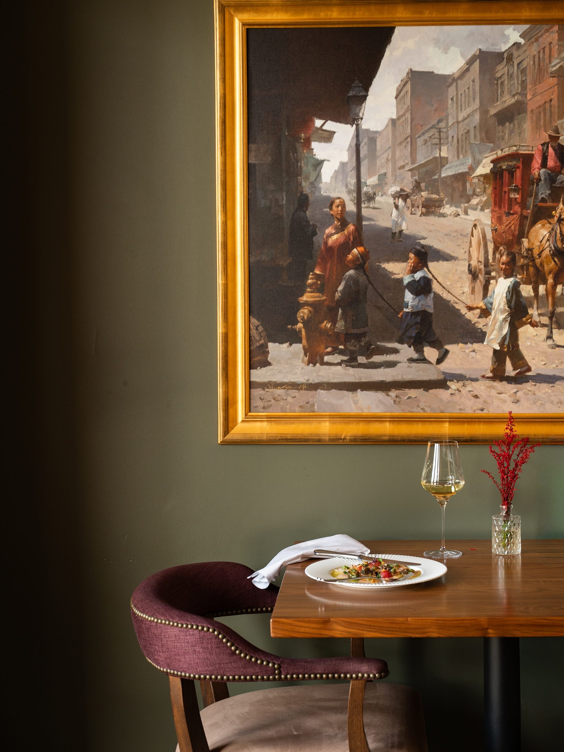 A restaurant table with food and wine sits beneath a framed street scene painting on a green wall, evoking the cozy elegance found at the Rusty Parrot Lodge and Spa.