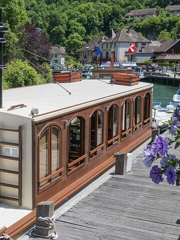 A vintage-style riverboat named "Le Chanaz," perfect for a luxury barge cruise, is docked on a calm canal with greenery and houses nearby.