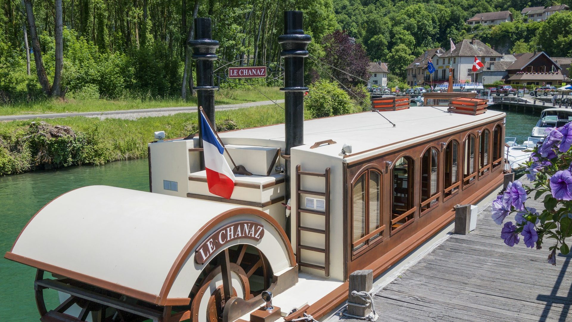 A vintage-style riverboat named "Le Chanaz," perfect for a luxury barge cruise, is docked on a calm canal with greenery and houses nearby.