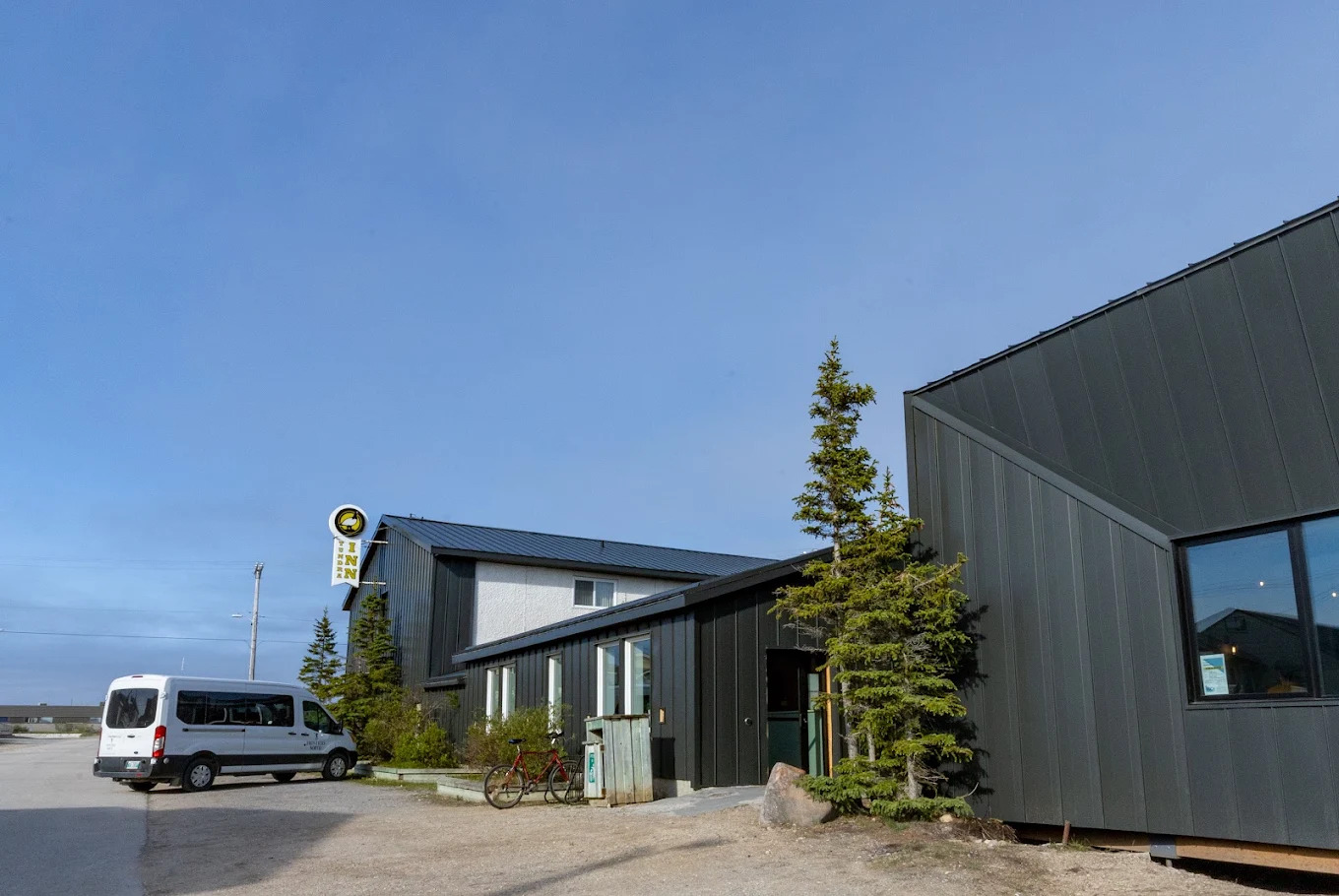 A white van parked next to a modern dark building with pine trees under a clear blue sky, ready to embark on an unforgettable Churchill Northern Lights tour.