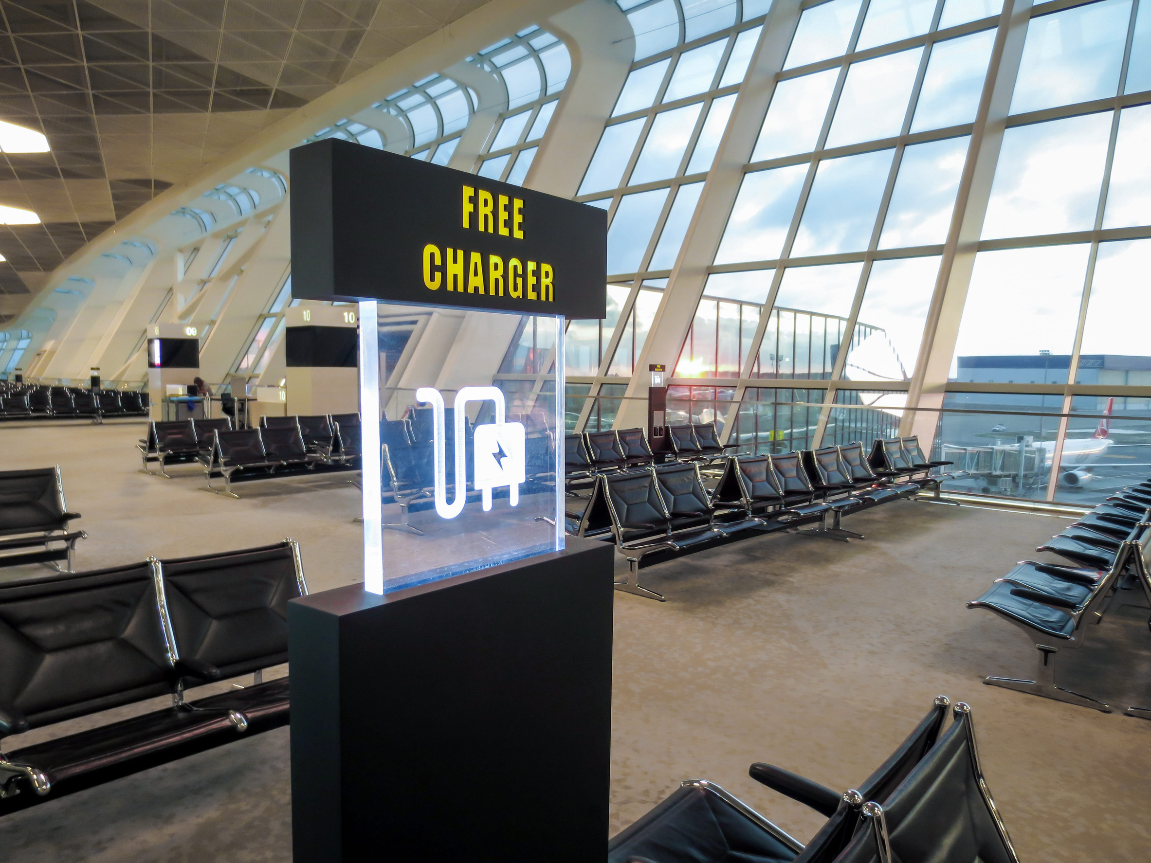 Airport seating area with large windows and a sign displaying “FREE CHARGER” and charging icons, highlighting convenient airport amenities for travelers.
