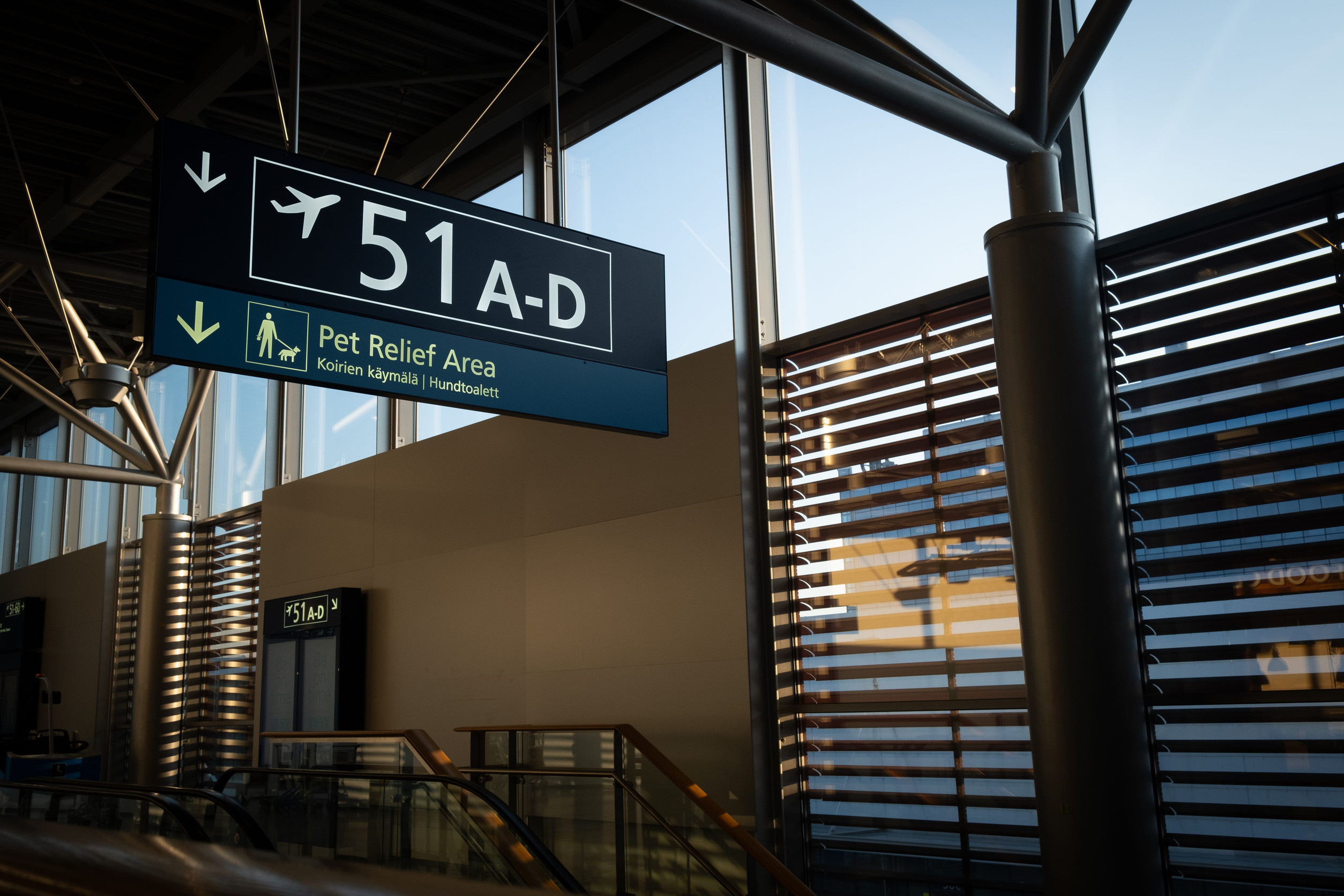 Airport sign for gates 51A-D and a pet relief area, showcasing essential airport amenities, with sunlight streaming through window slats.