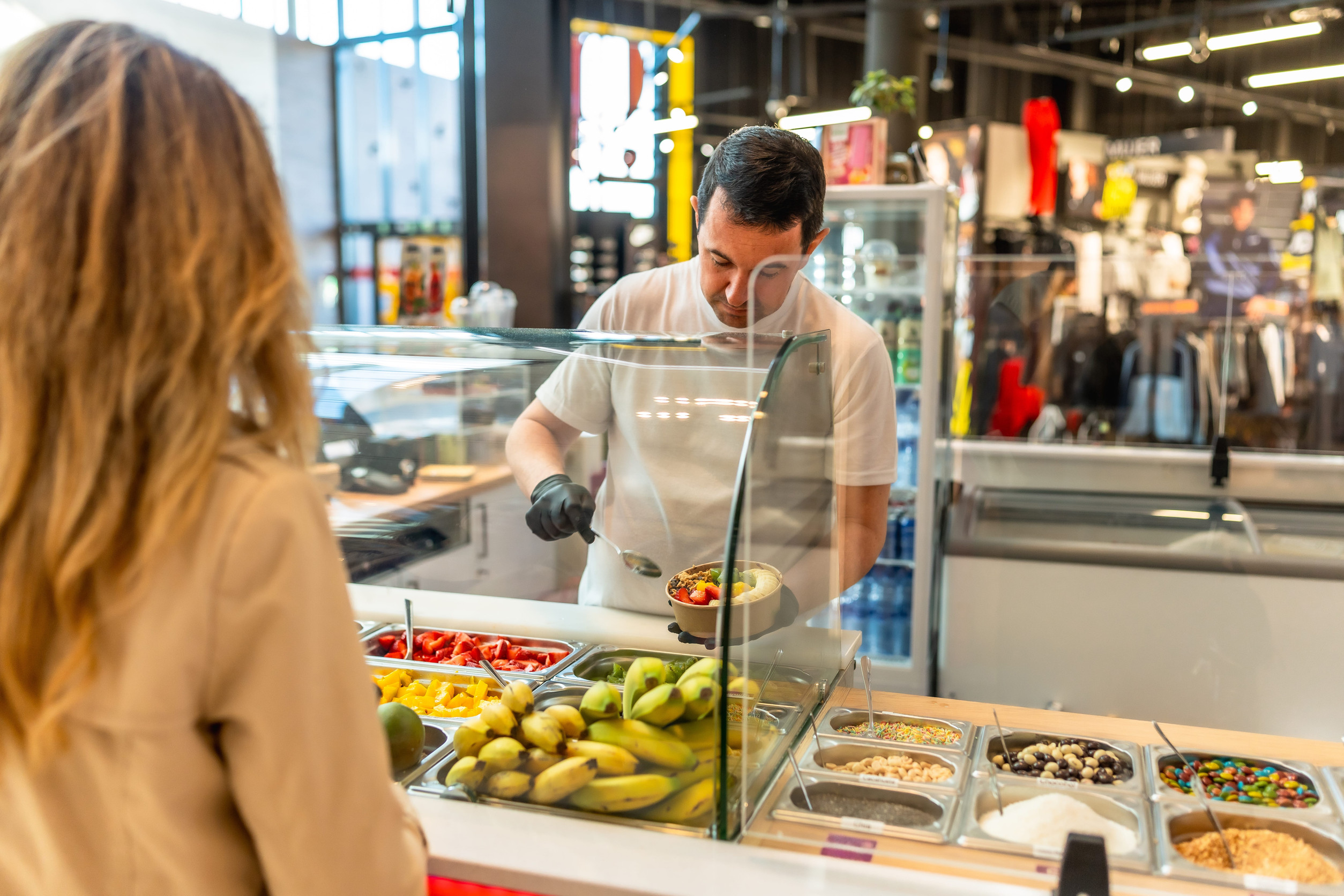A worker prepares a fruit bowl behind glass as a customer waits at a counter in a modern food shop, highlighting the convenient airport amenities that cater to travelers seeking fresh and healthy options.