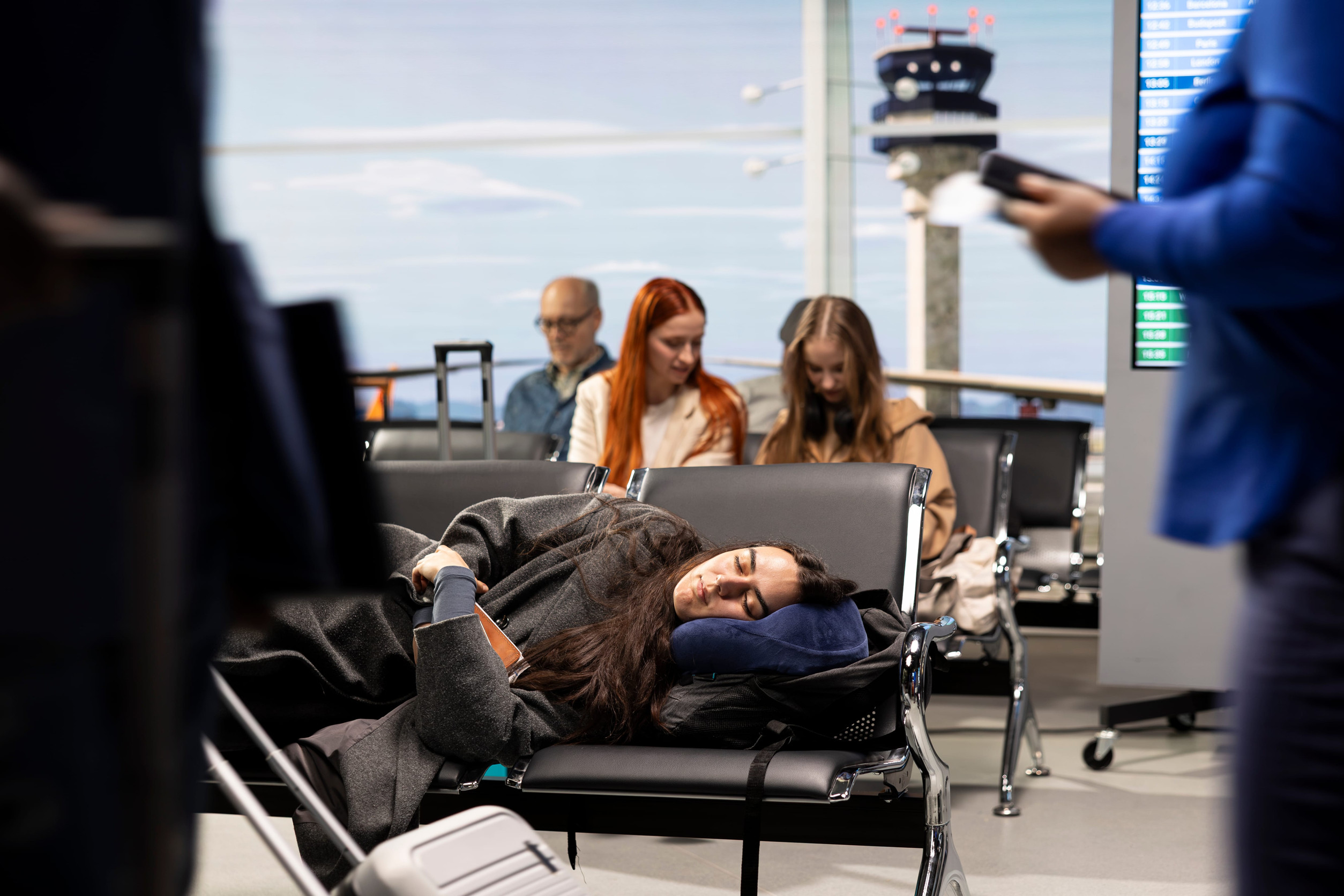 Woman sleeping on airport seats with luggage; people sitting and checking phones in the background, making use of available airport amenities.
