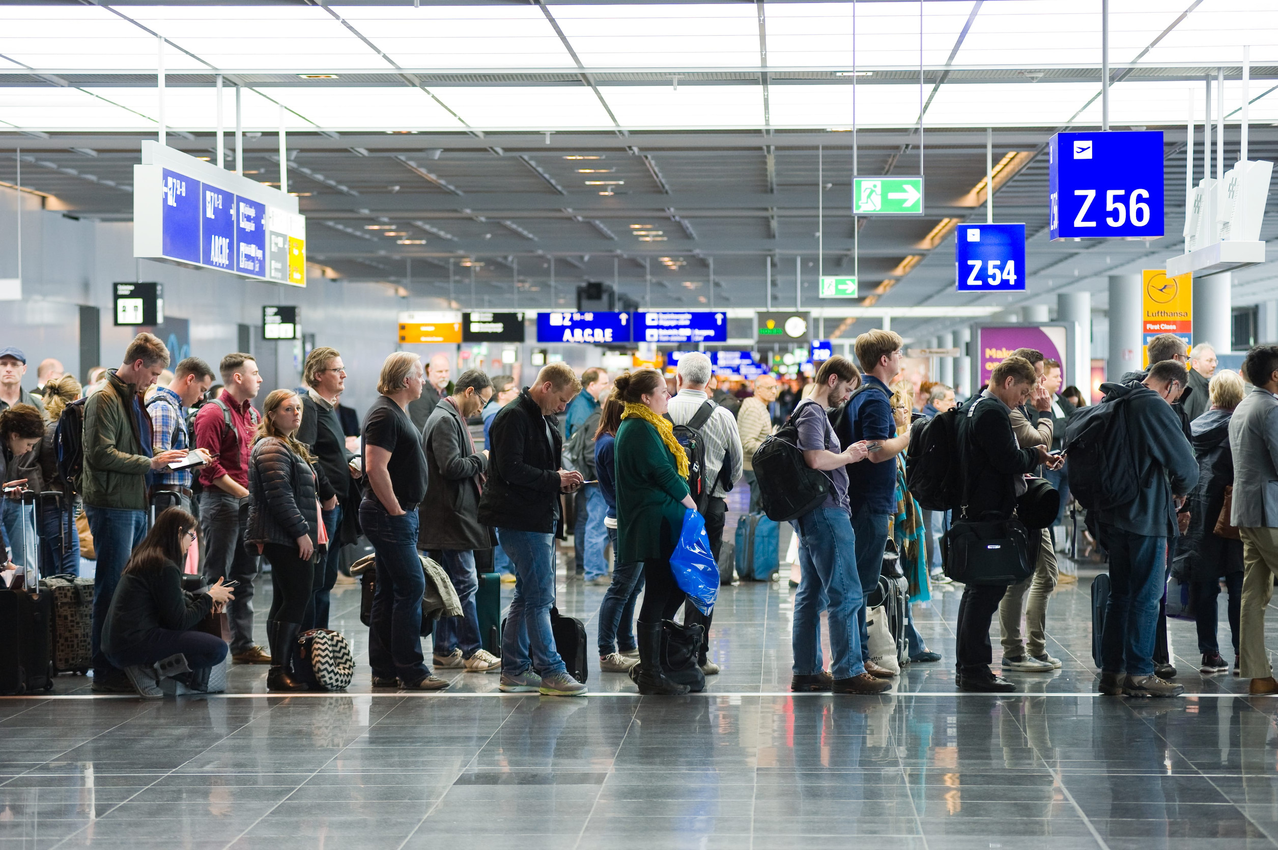Travelers wait in a long line at an airport terminal under bright lights and blue gate signs, anticipating the comfort of convenient airport amenities ahead.