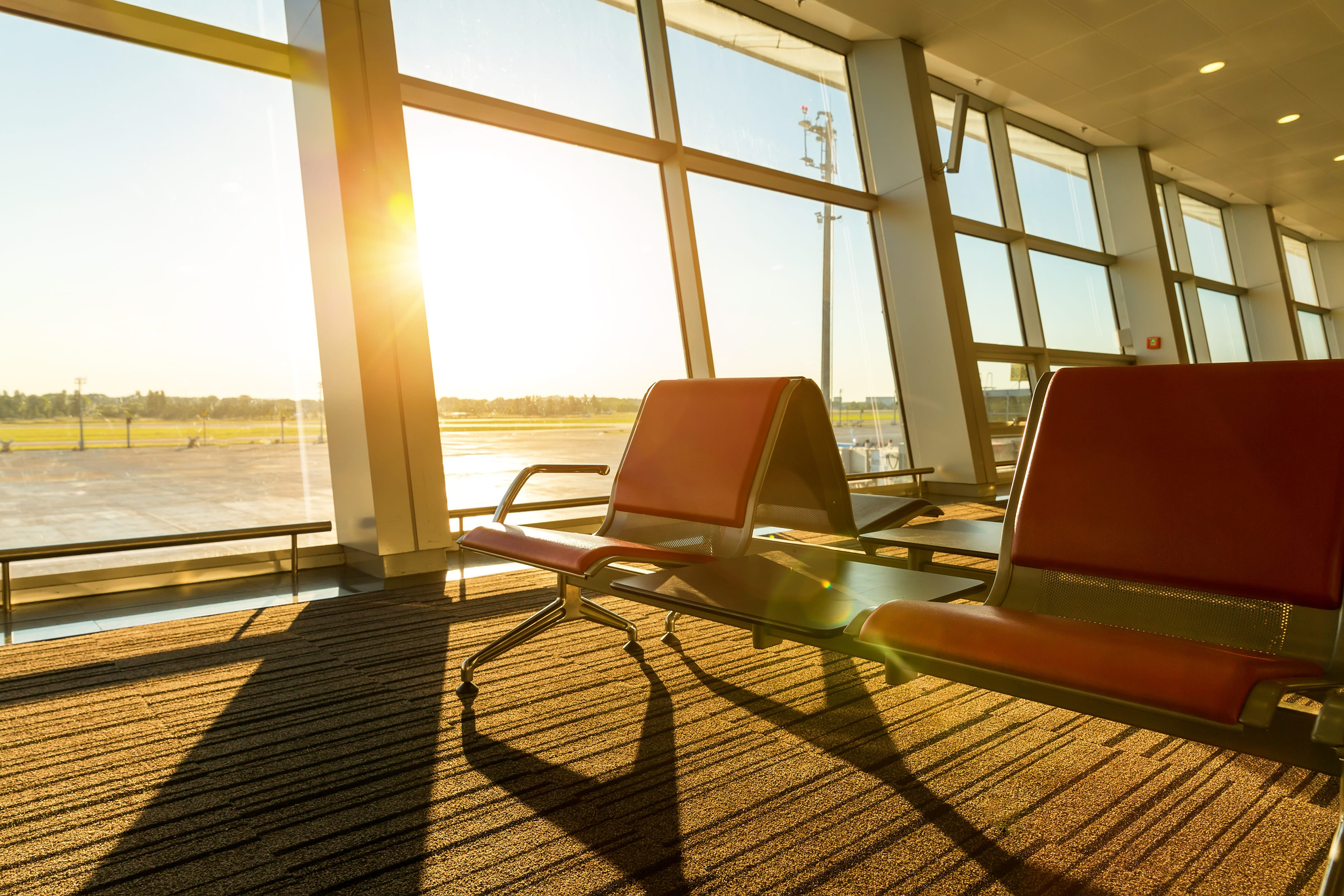 Sunlight streams through large airport windows, illuminating empty red chairs in the waiting area, a tranquil space enhanced by thoughtful airport amenities.