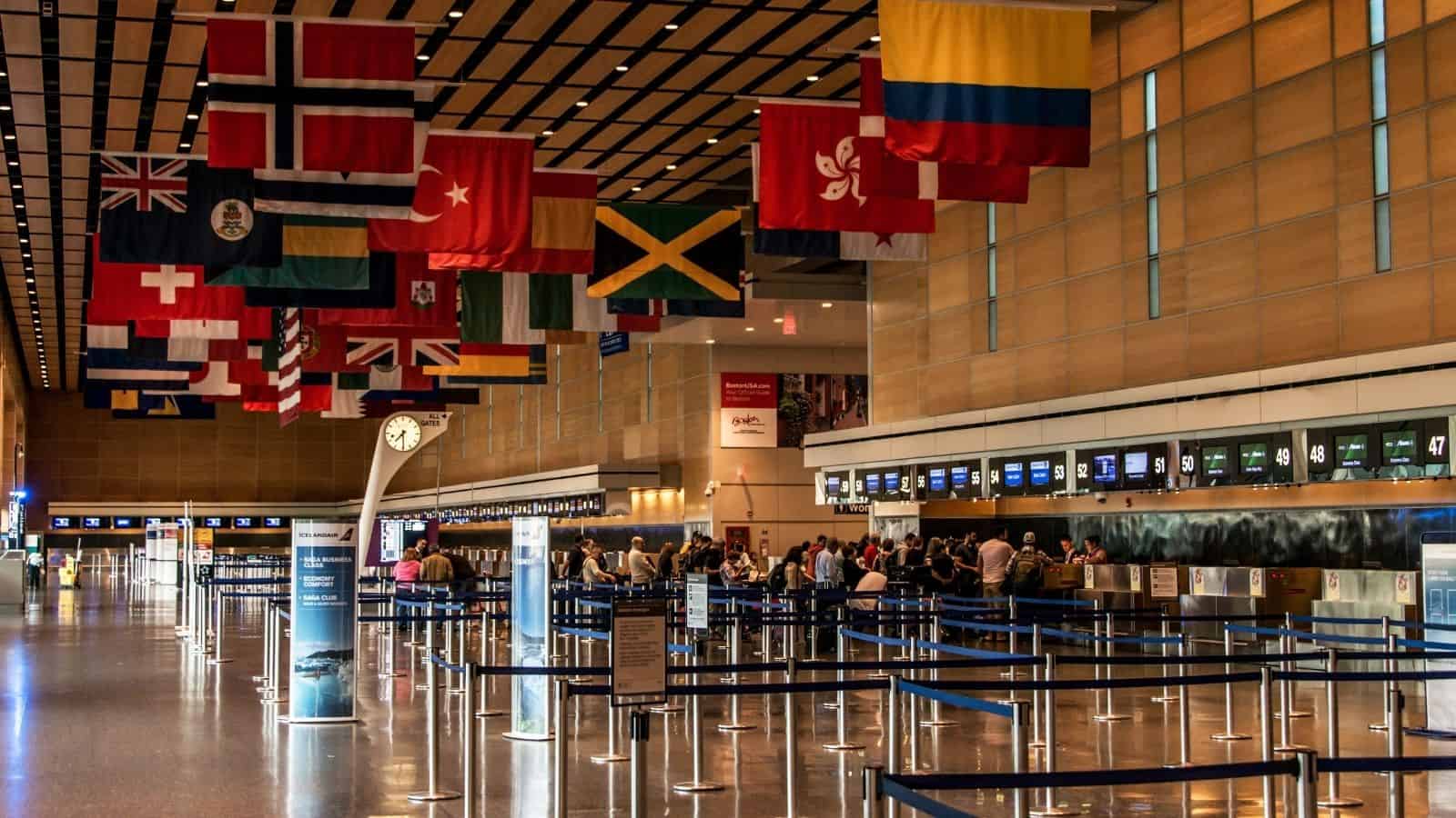 Airport check-in area with international flags hanging above, people waiting in lines, and empty lanes.