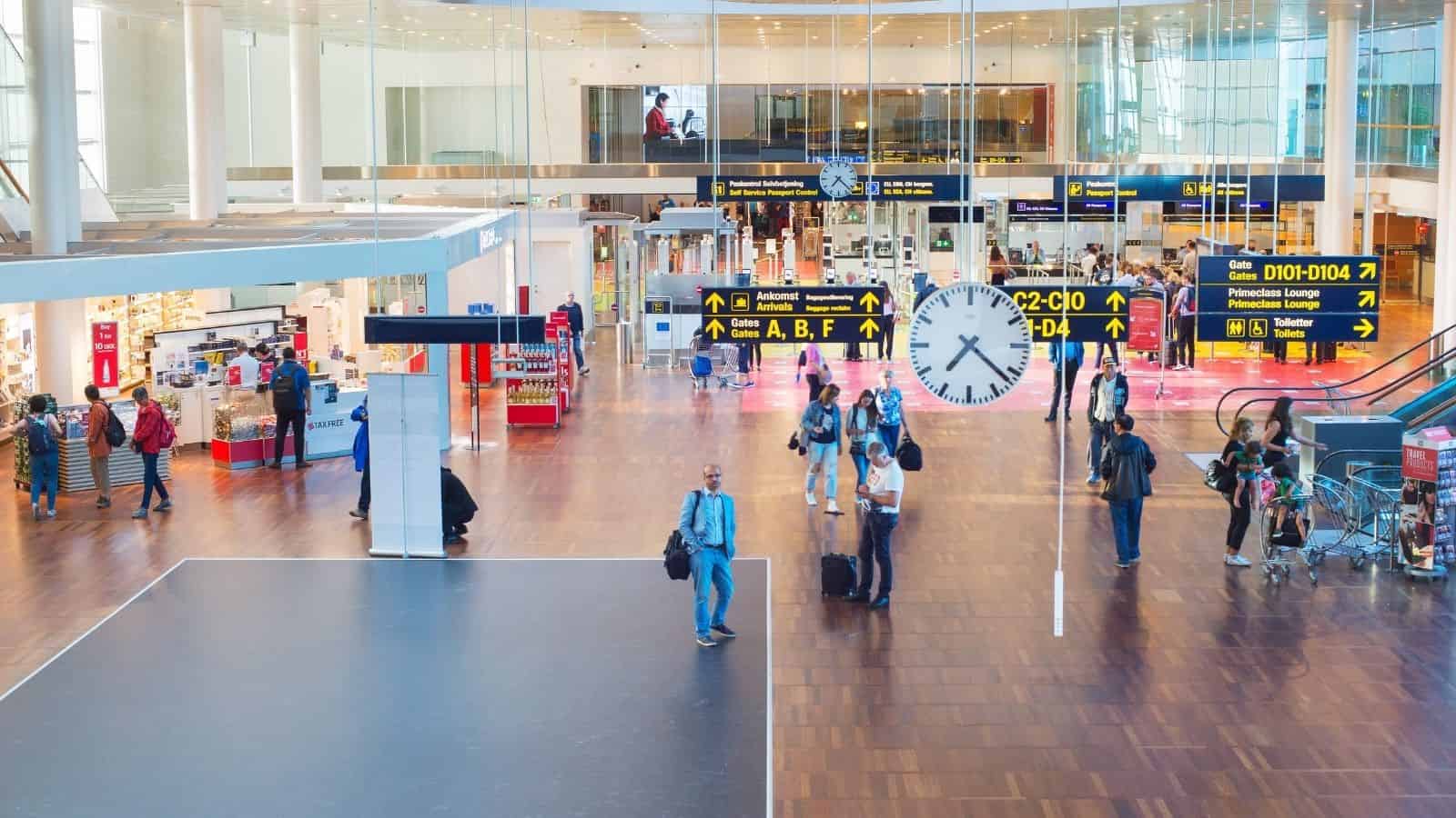 People walk through a bright airport terminal with shops, signs, and a large clock in the center.