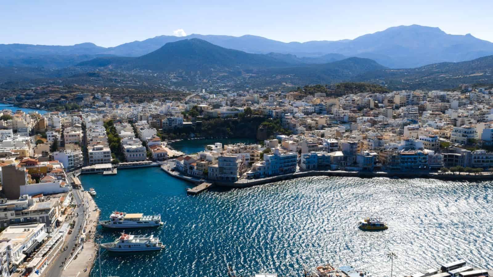 A coastal city with white buildings, boats in the harbor, and mountains in the background under a clear sky.