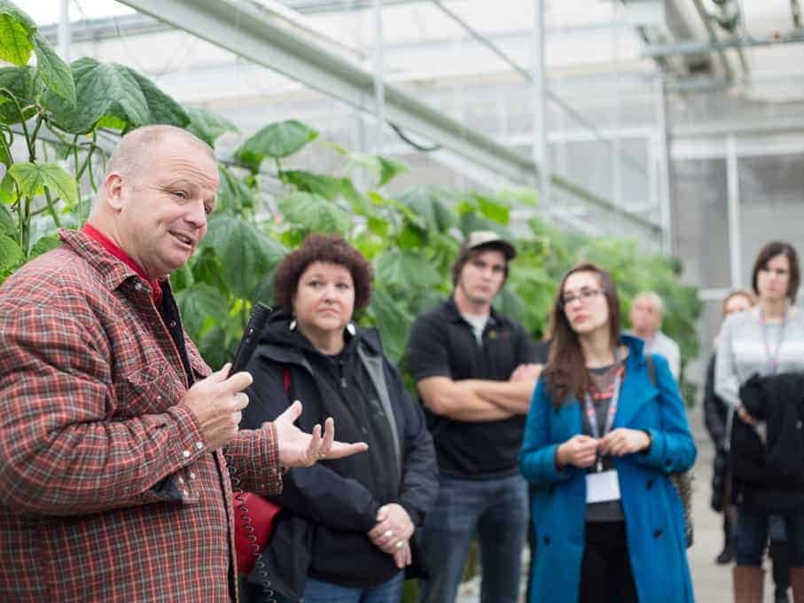 A man speaks to a group of people in a greenhouse with green plants in the background, sharing insights about sustainable practices from Farm and Food Care Ontario.
