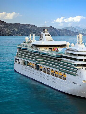A large white cruise ship sails on calm blue water with mountains and a clear sky in the background, offering guests a front-row seat to an unforgettable Solar Eclipse Cruise experience.