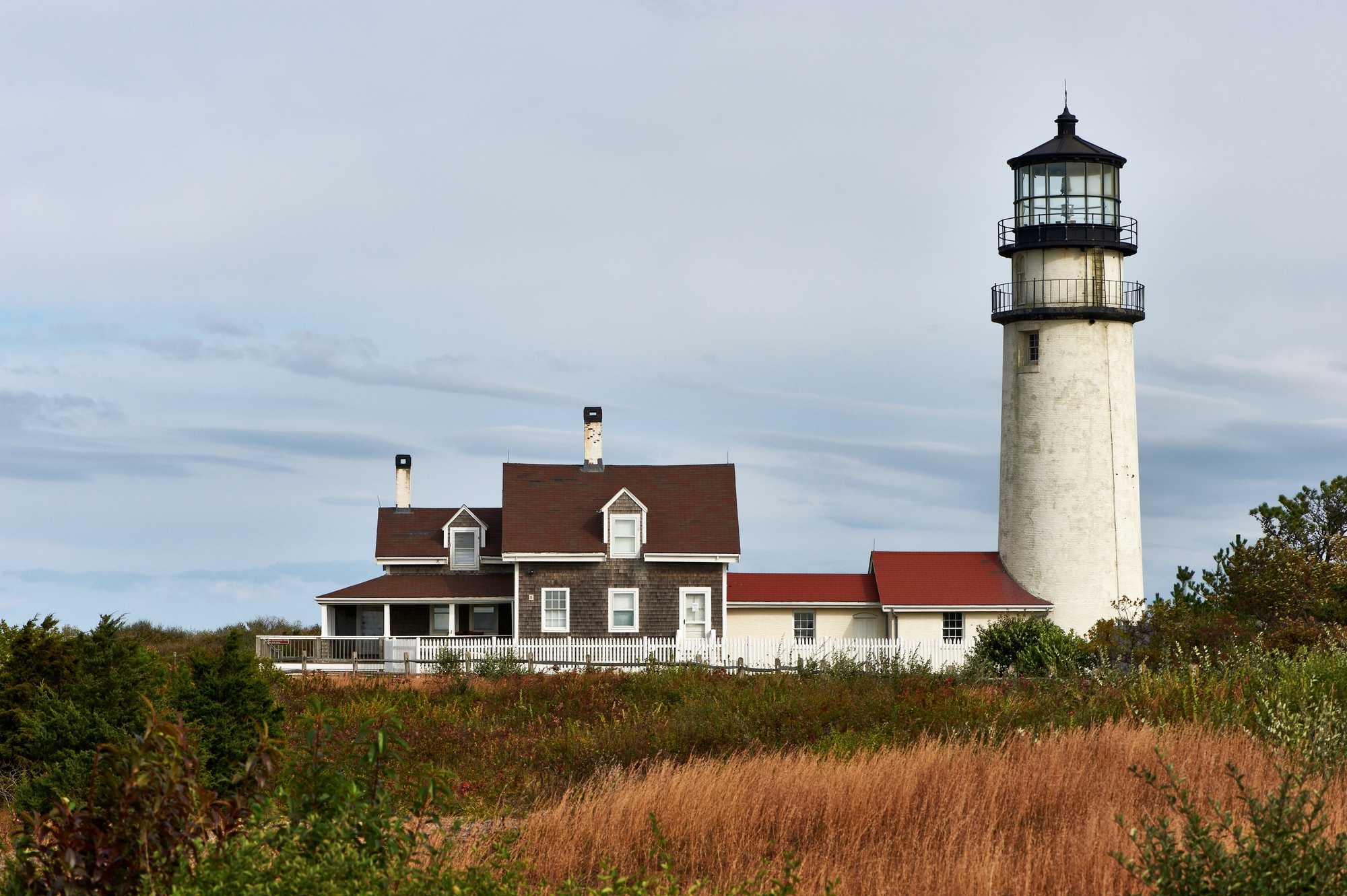 A tall white lighthouse with an attached house, surrounded by grass and shrubs under a cloudy sky, evokes the charm of european destination dupes.