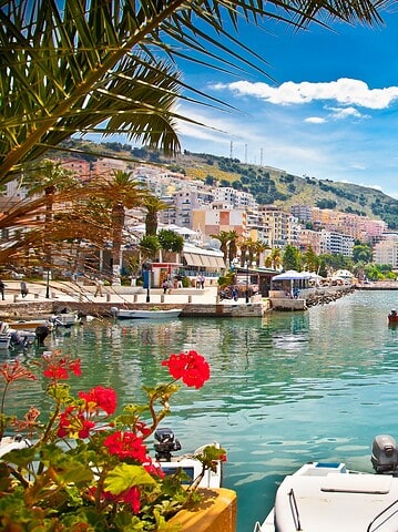 Colorful boats docked in a marina, framed by palm leaves and red flowers, with a city and hills in the background—this picturesque setting is one of the top European destination dupes, offering all the charm of a classic coastal getaway without the crowds.