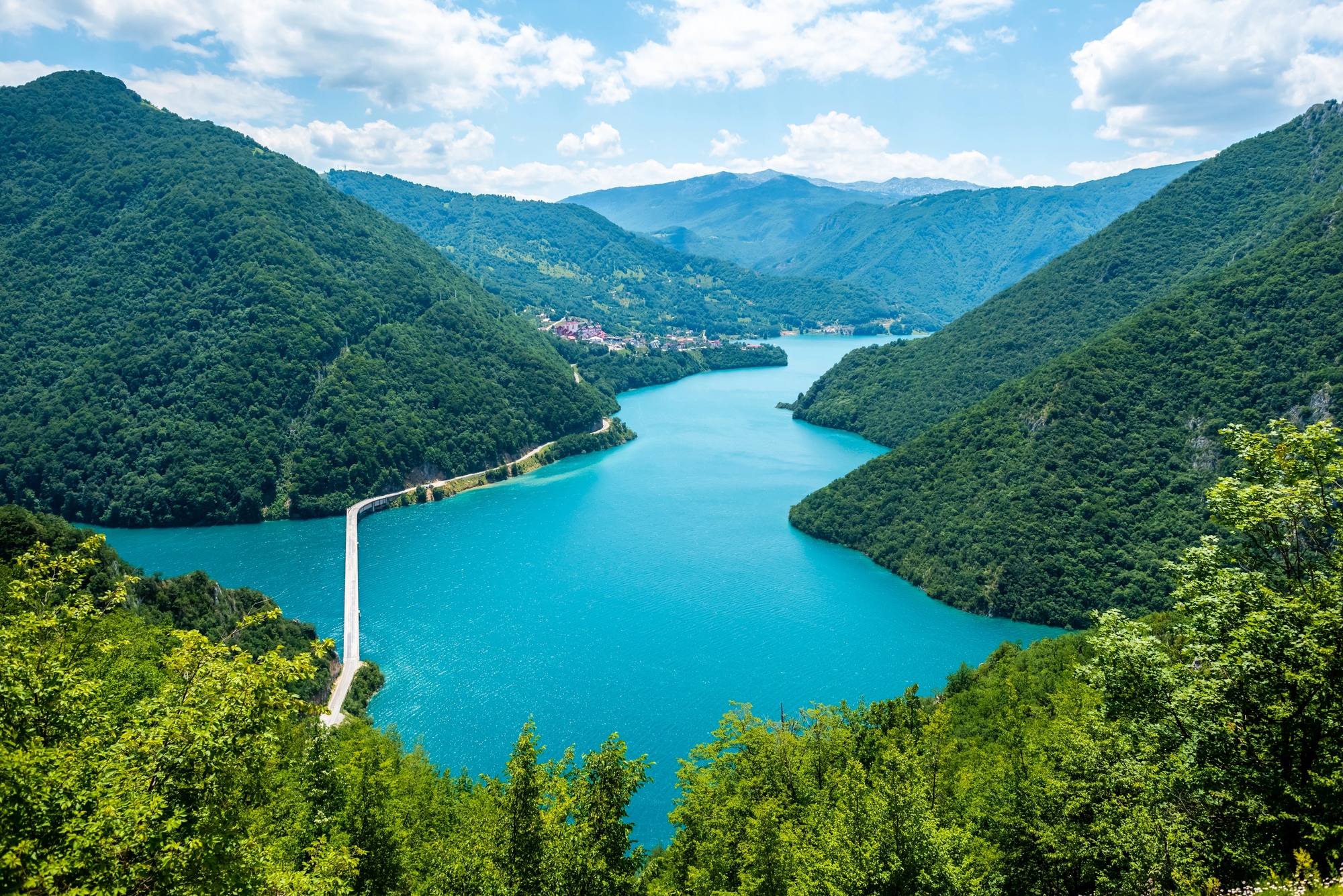 A winding river flows between lush, green hills under a bright, partly cloudy sky, evoking the charm of hidden European destination dupes waiting to be discovered.