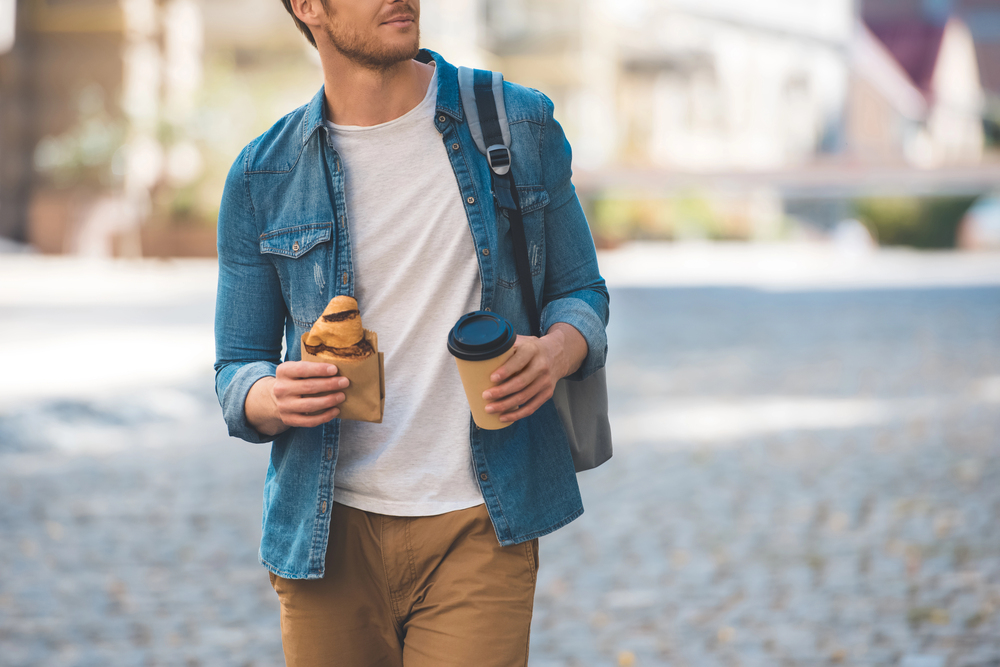 Man in casual clothes holding a sandwich and a coffee cup while walking outdoors, enjoying what could be the start of a delicious food tour.