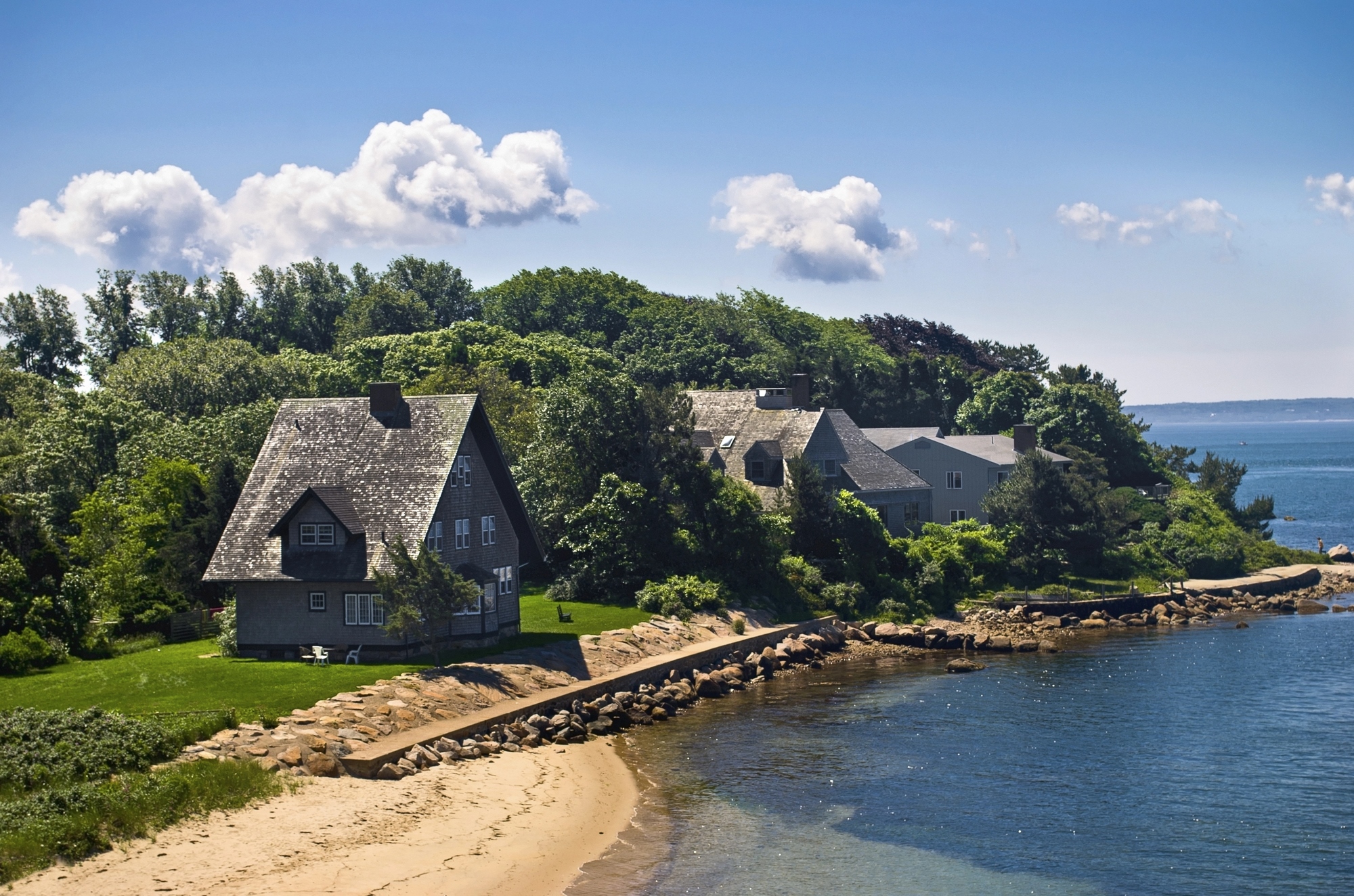 Gray seaside cottages surrounded by trees along a sandy shoreline under a bright blue sky, this hidden gem is one of those European destination dupes perfect for a tranquil getaway.