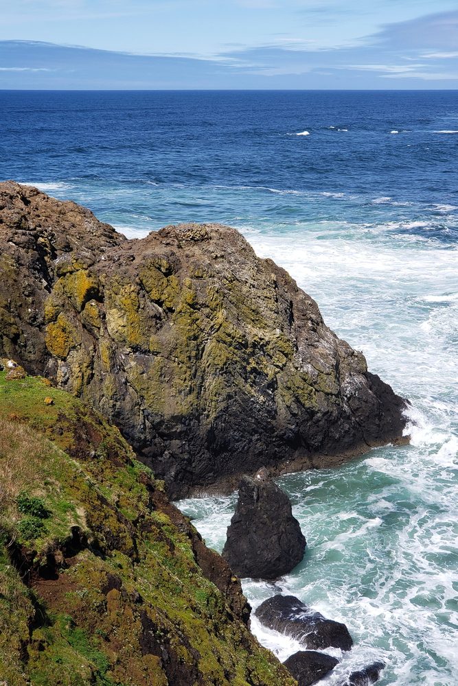 Rocky coastline with waves crashing against mossy cliffs under a partly cloudy blue sky&mdash;an idyllic scene reminiscent of hidden European destination dupes waiting to be explored.