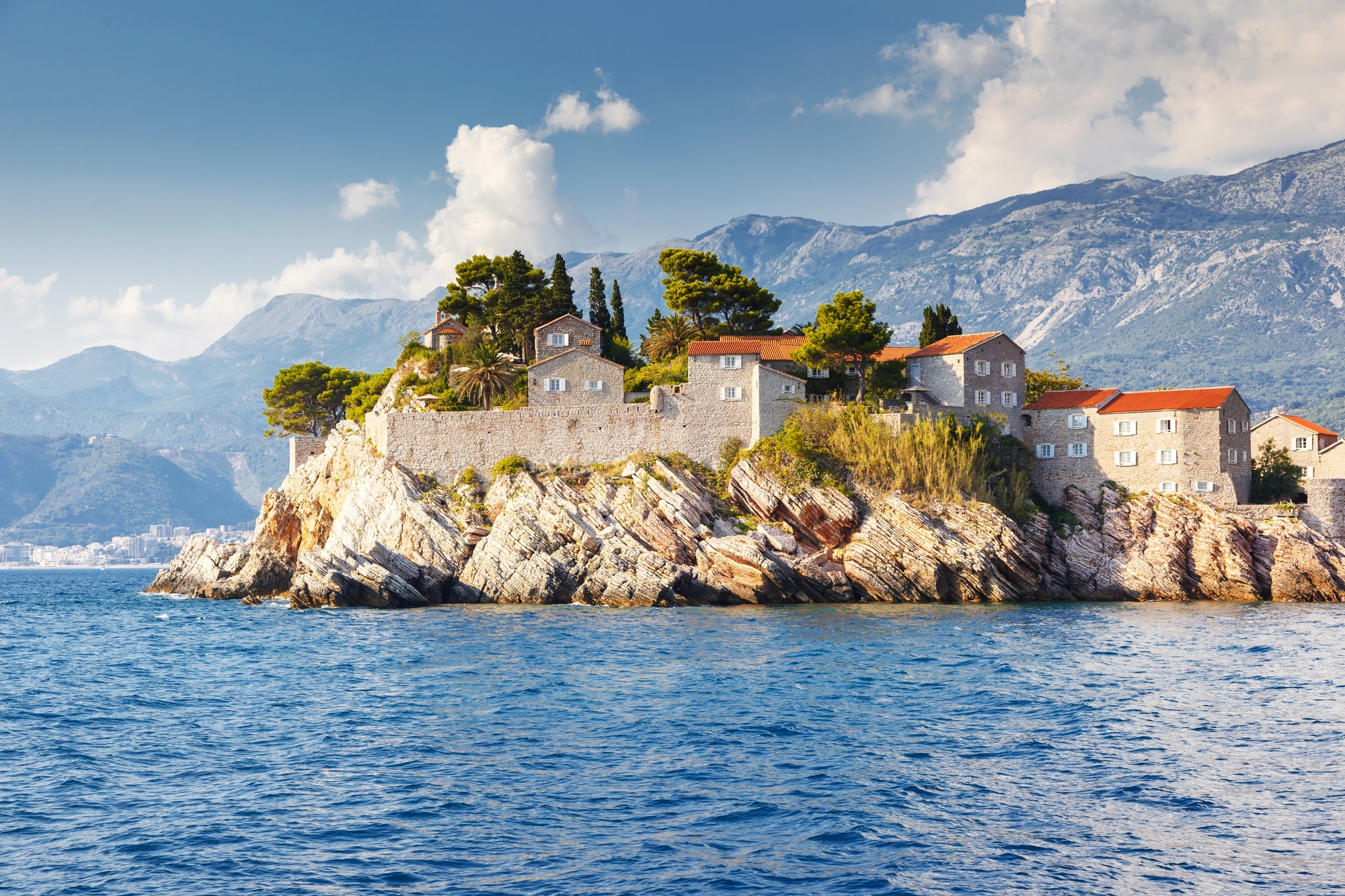 Stone buildings with red roofs perch on a rocky island surrounded by blue sea and distant mountains, evoking the charm of sought-after European destination dupes.