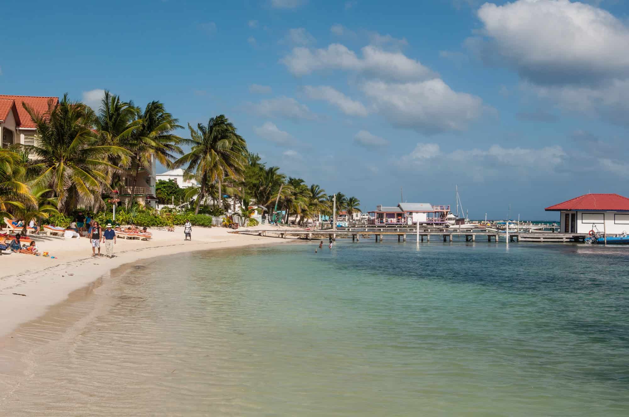 Sandy beach with palm trees, people walking, and docks extending into clear, blue-green water under a sunny sky&mdash;the perfect starting point for your Belize reef and ruins trip.