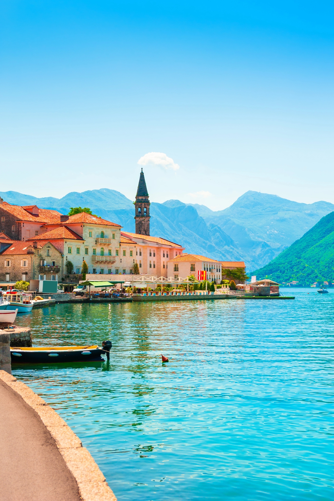 Colorful coastal village with mountains, boats on blue water, and a church tower under a clear sky&mdash;perfect for travelers seeking scenic European destination dupes.