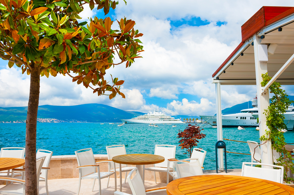 Outdoor caf&eacute; by the sea with empty tables, a tree, yachts, and mountains under a blue sky with clouds&mdash;capturing the charm of european destination dupes for those seeking a picturesque escape without leaving home.