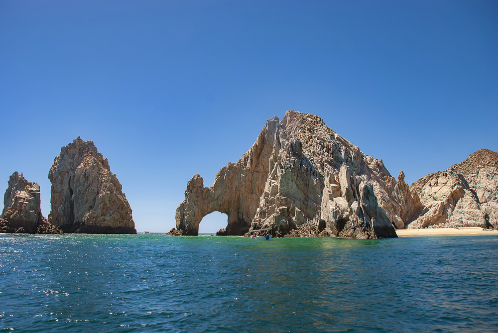Rock arch formations rise from turquoise sea under a clear blue sky near a sandy shore, creating the perfect scene for travelers seeking stunning European destination dupes.