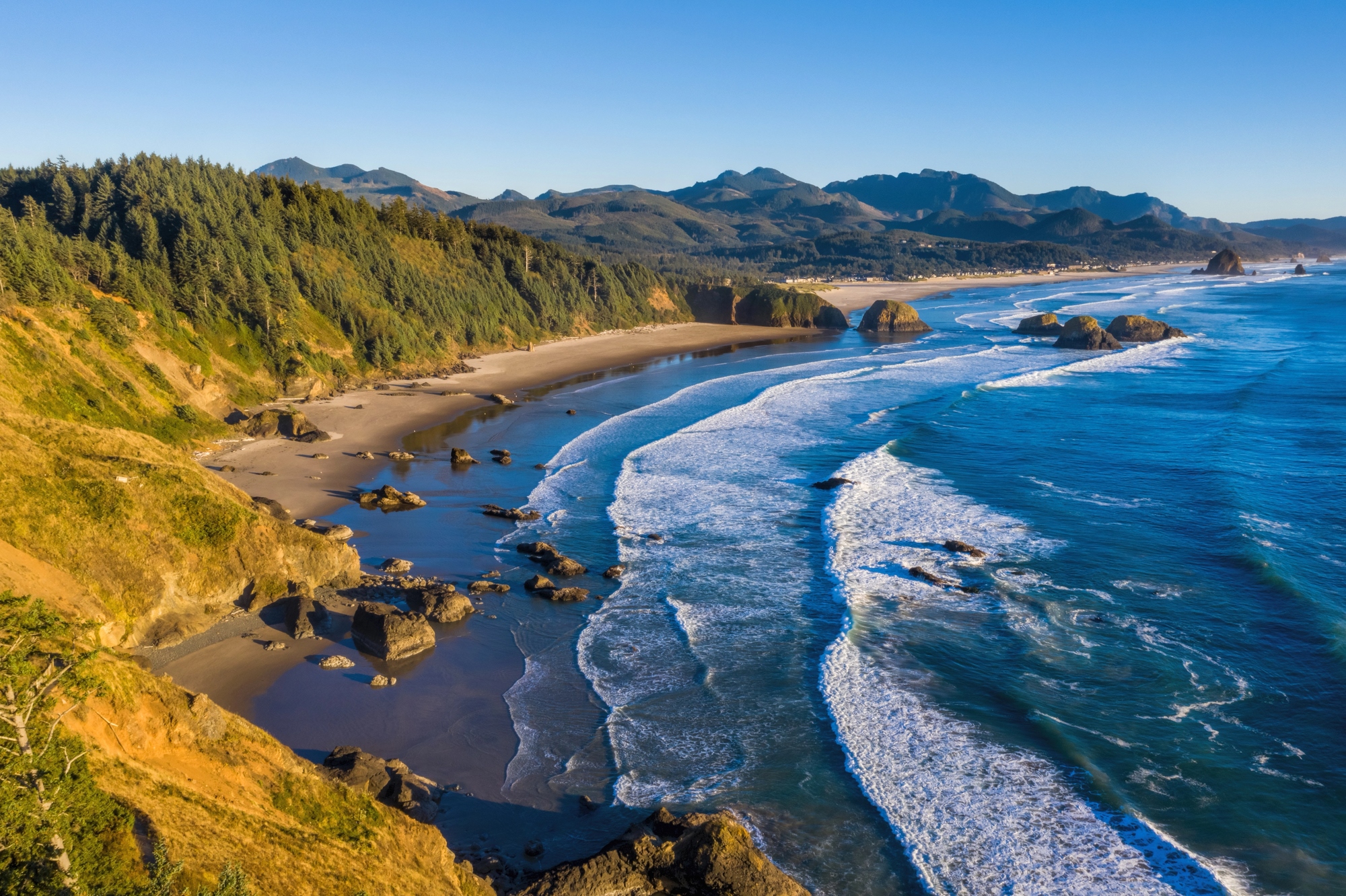 Rocky coast with waves, sandy beach, forested cliffs, and distant mountains under a clear blue sky&mdash;this tranquil setting offers the charm of European destination dupes without the crowds.