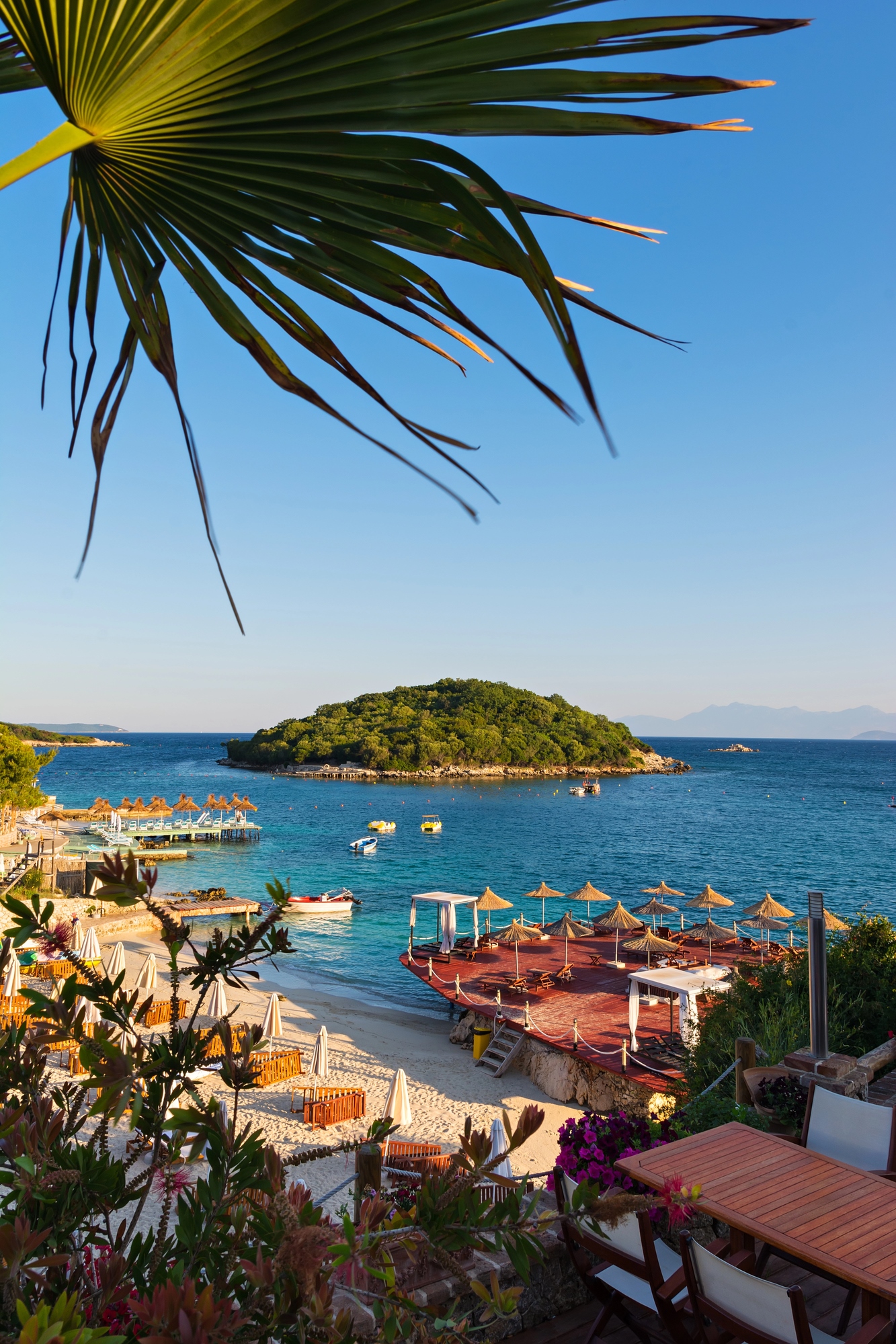 Sunny beach with umbrellas, lounge chairs, and boats, overlooking a small green island in blue water&mdash;a perfect spot for travelers seeking european destination dupes without the crowds.