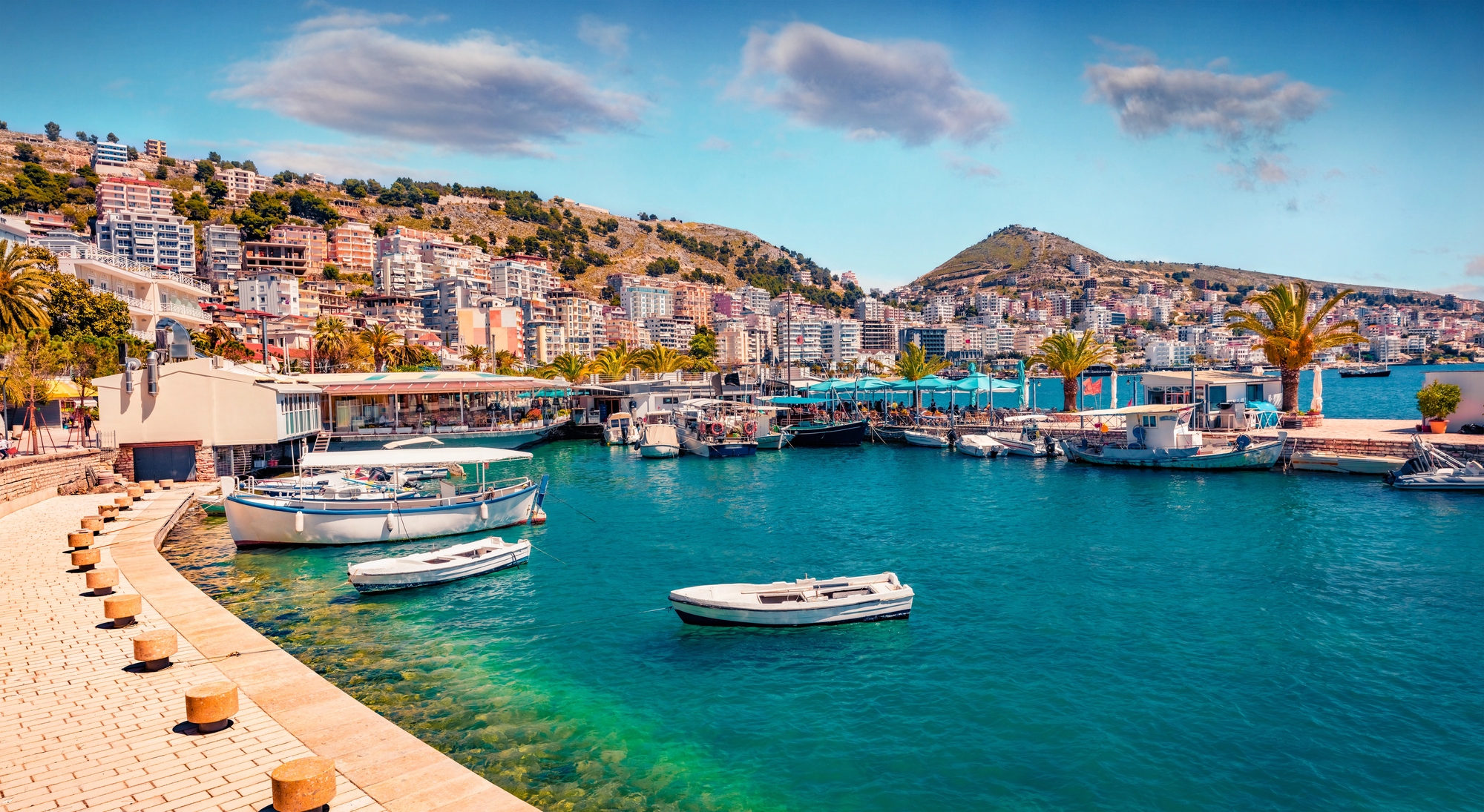 Boats docked in a turquoise harbor, with buildings and hills in the background under a partly cloudy sky&mdash;an idyllic scene reminiscent of popular European destination dupes.
